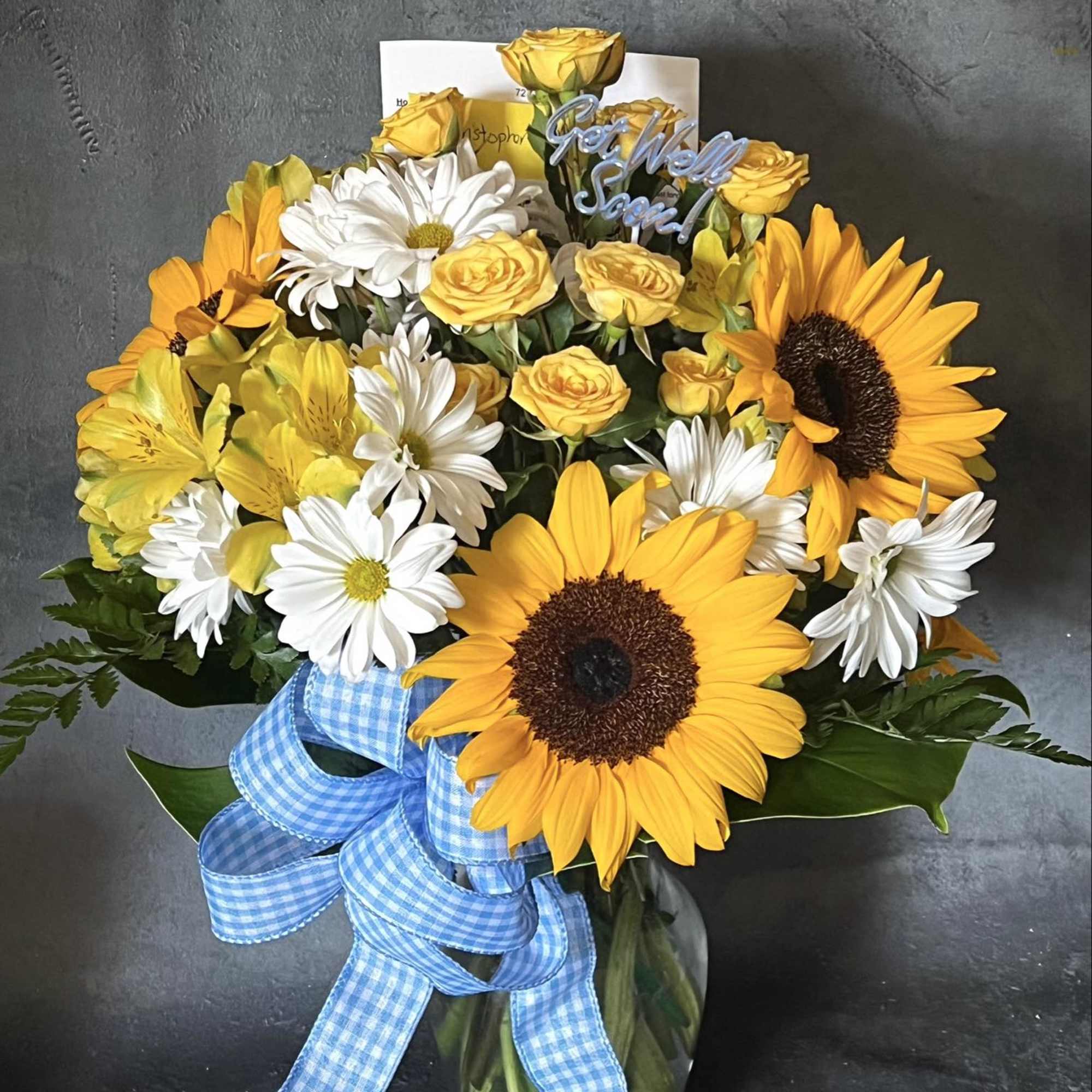 Bouquet of yellow roses, sunflowers, and white daisies in a glass vase with blue ribbon