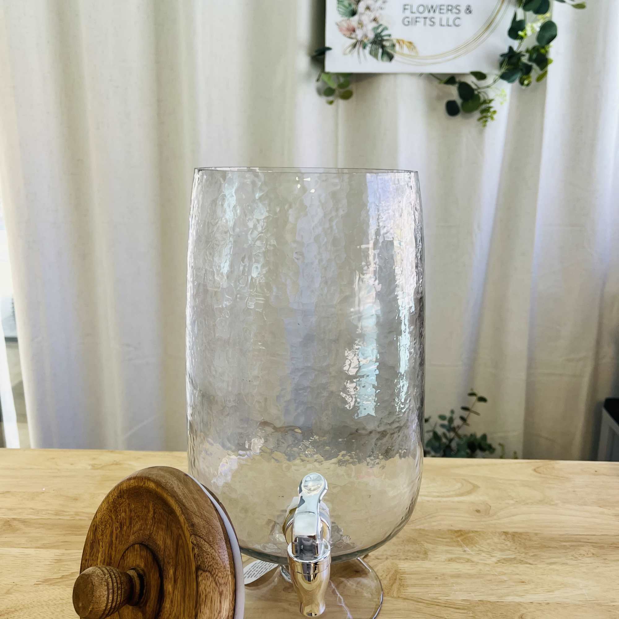 Hammered glass drink dispenser with metal spout and wooden lid on a light wood surface