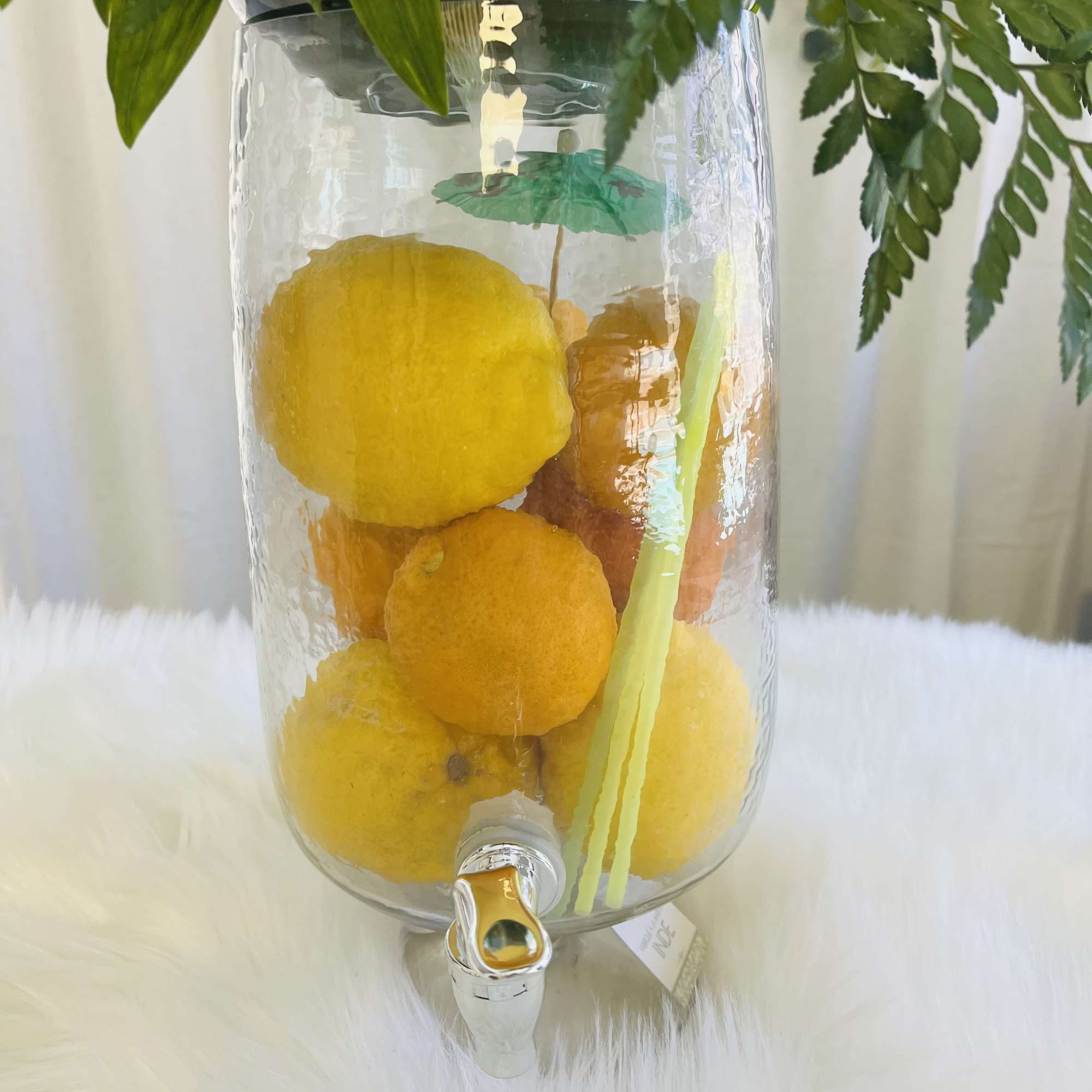 Glass drink dispenser filled with lemons beneath yellow daisy flowers