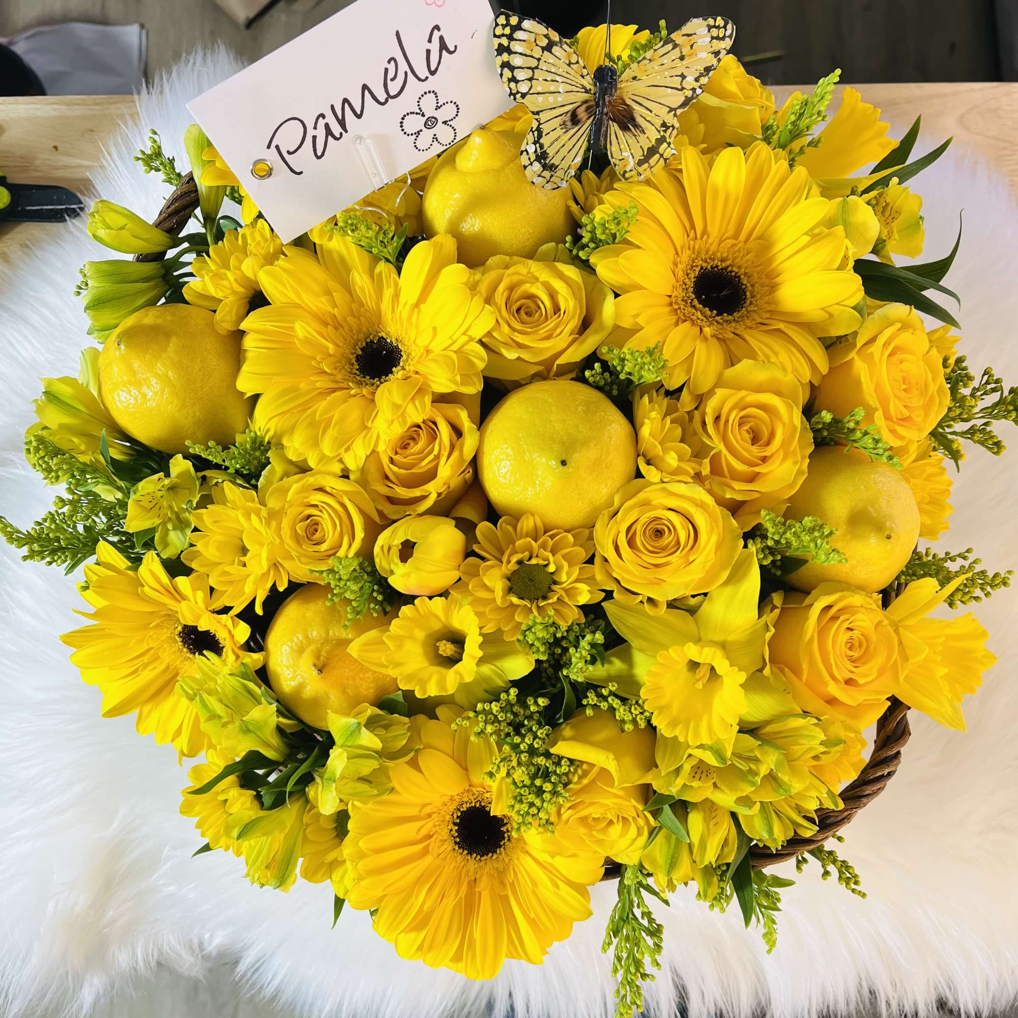 Round yellow flower basket with gerbera daisies, roses, lemons, and a name card topped with a butterfly.