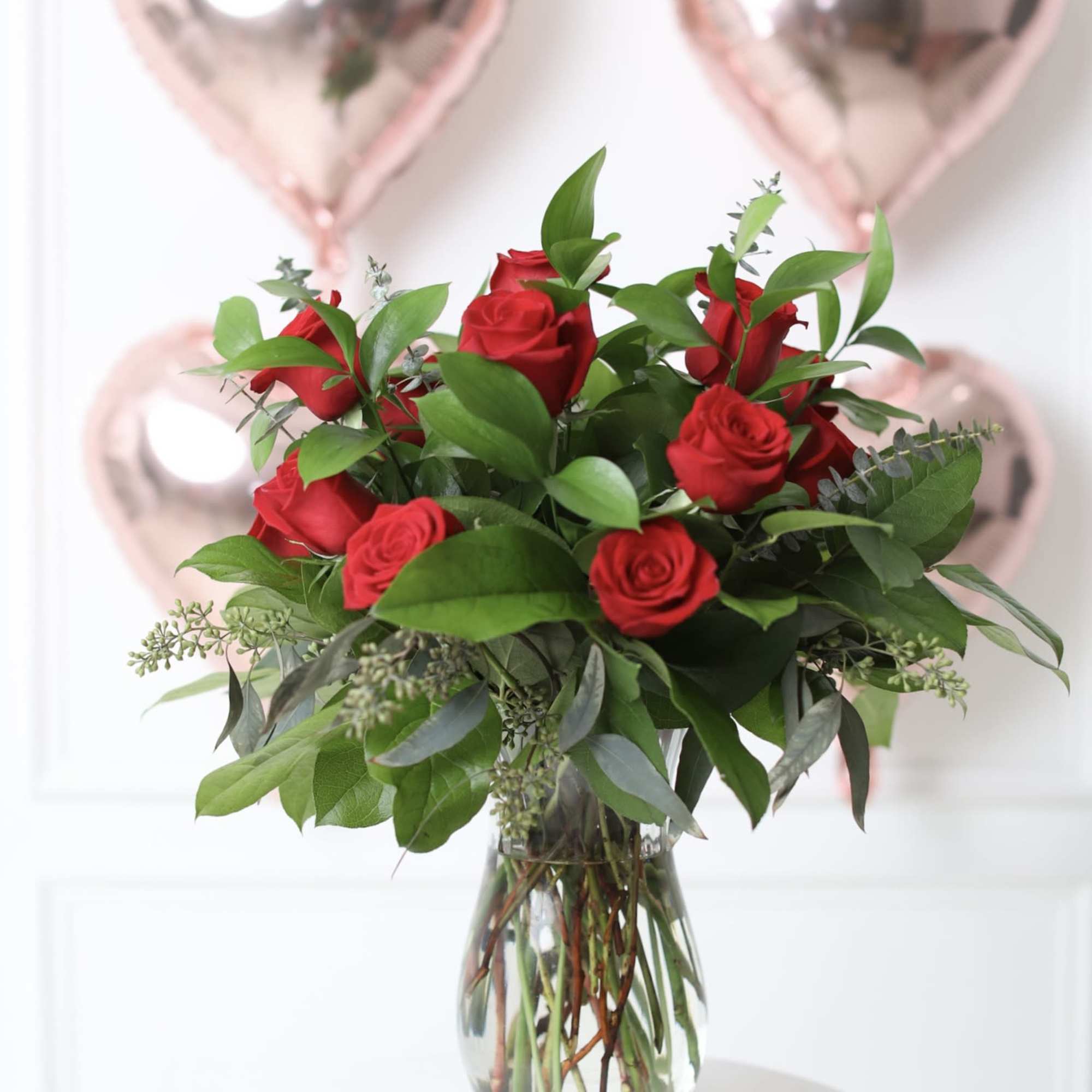 Red roses in a clear glass vase with pink heart balloons behind them
