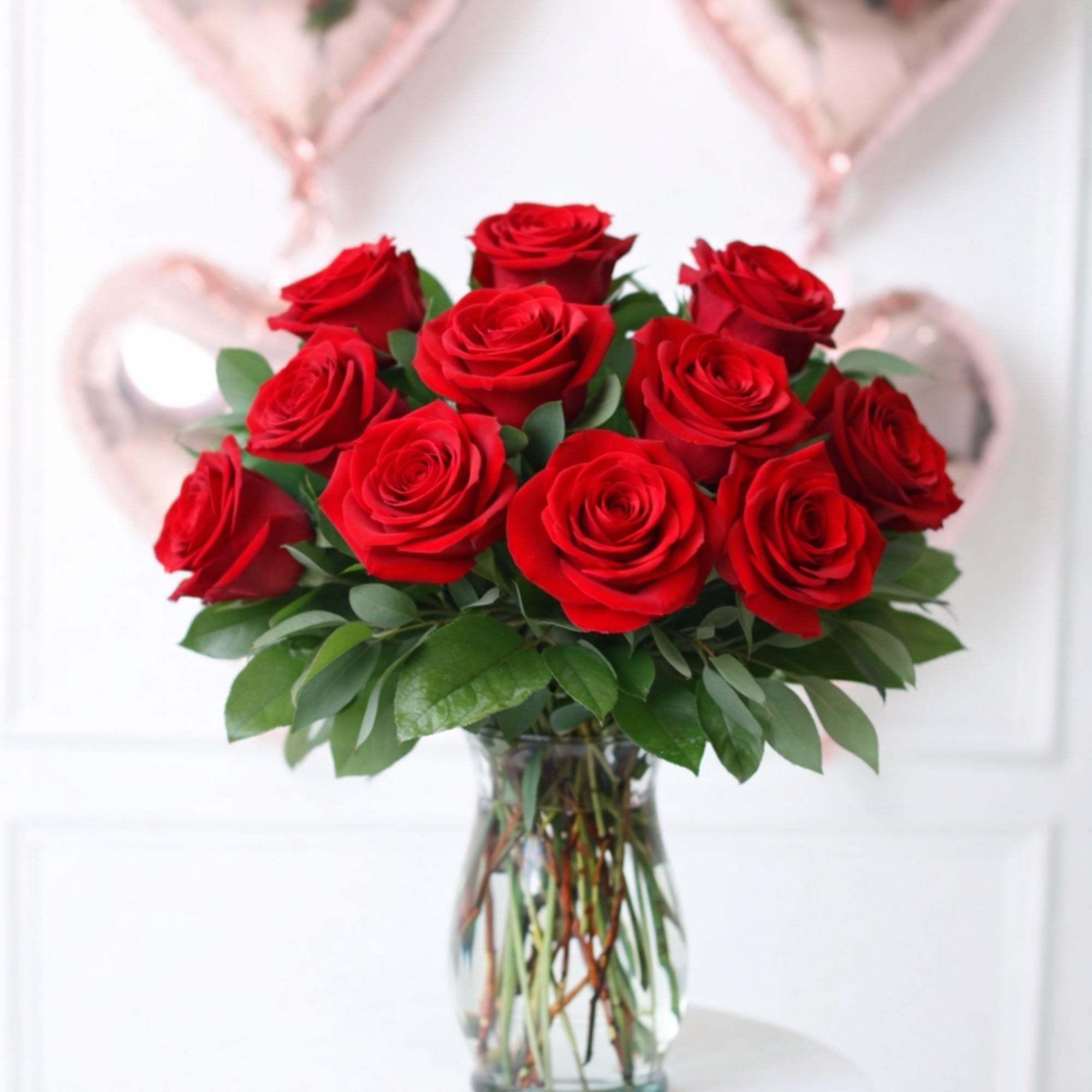 Red roses in a clear glass vase with pink heart balloons behind