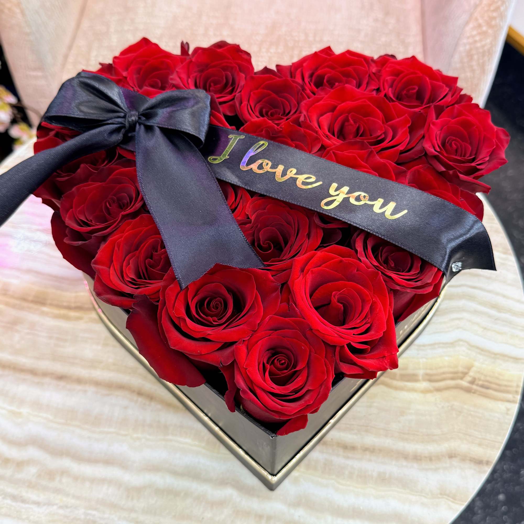 Heart-shaped box of red roses with a black ribbon reading "I love you"