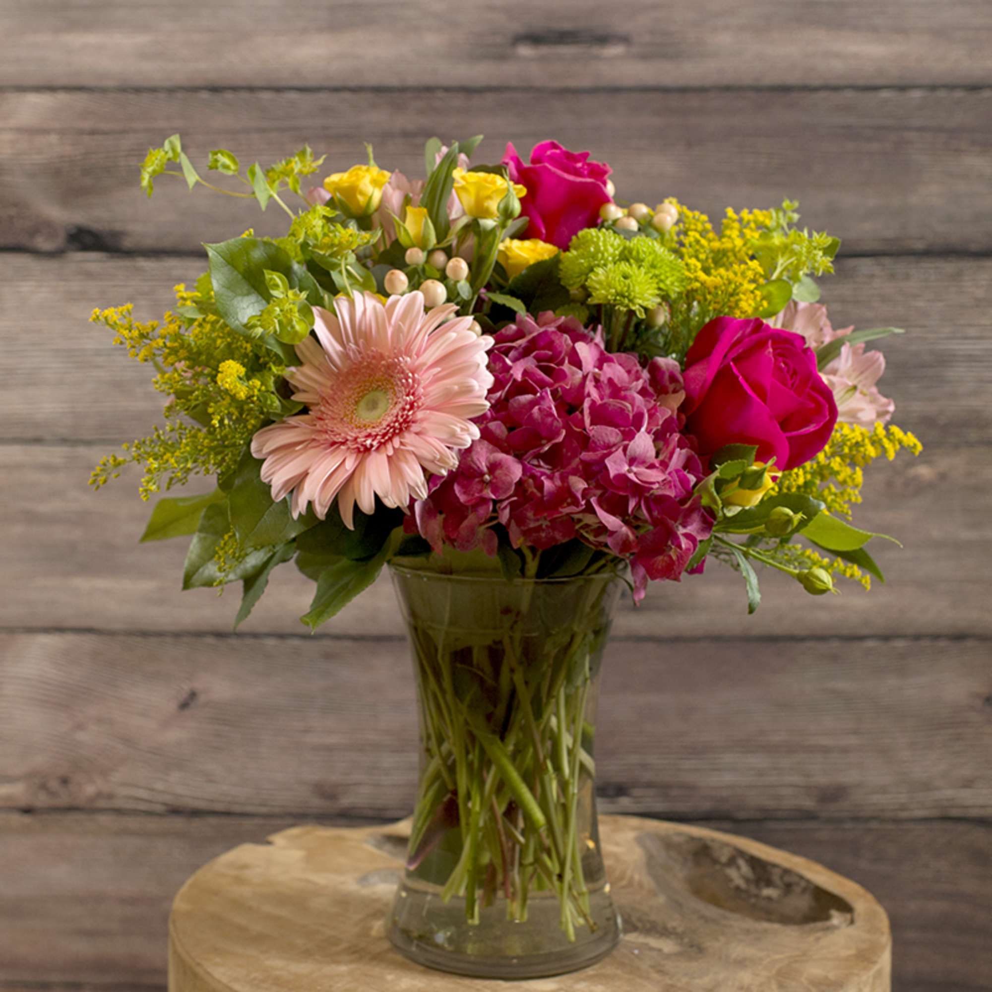 Mixed bouquet of pink roses, gerbera daisy, and hydrangea in a clear glass vase on a wooden table