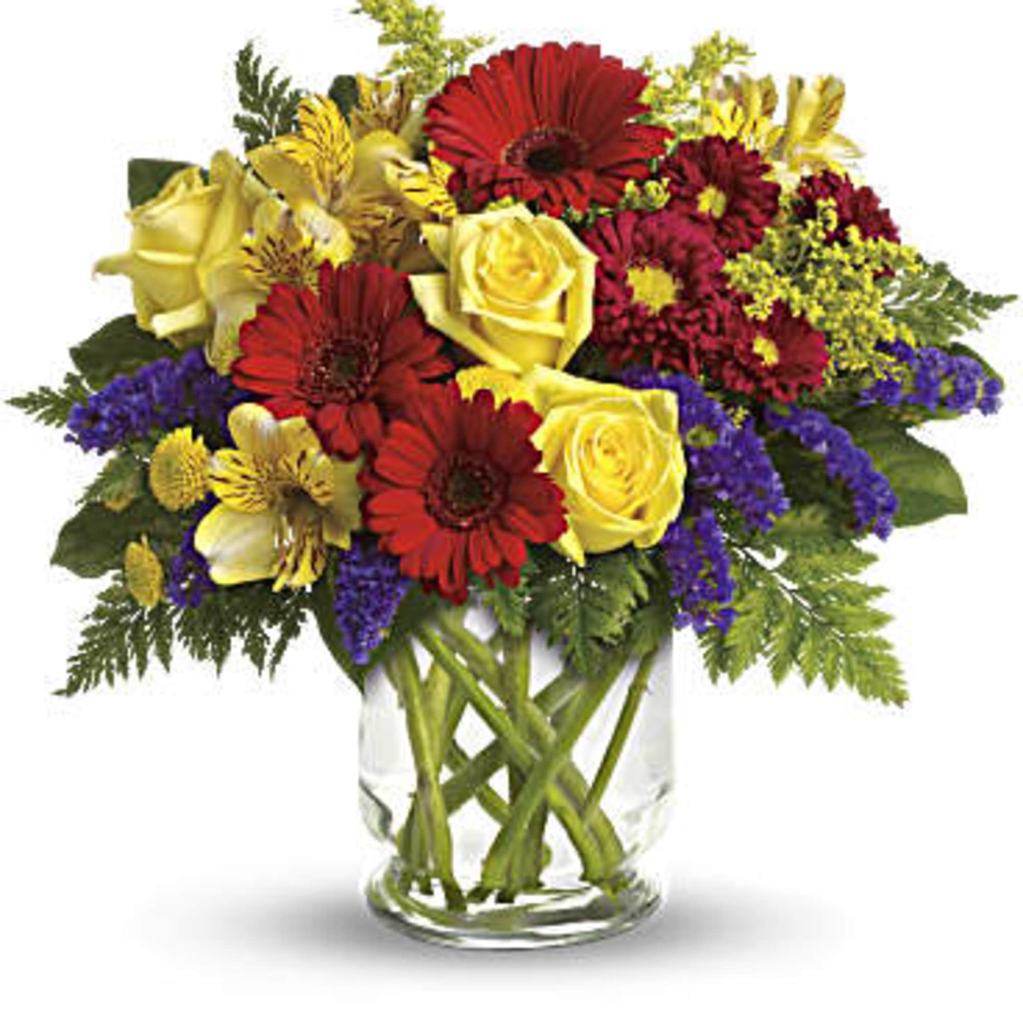 Bright arrangement of red gerbera daisies, yellow roses, and purple blooms in a clear glass vase