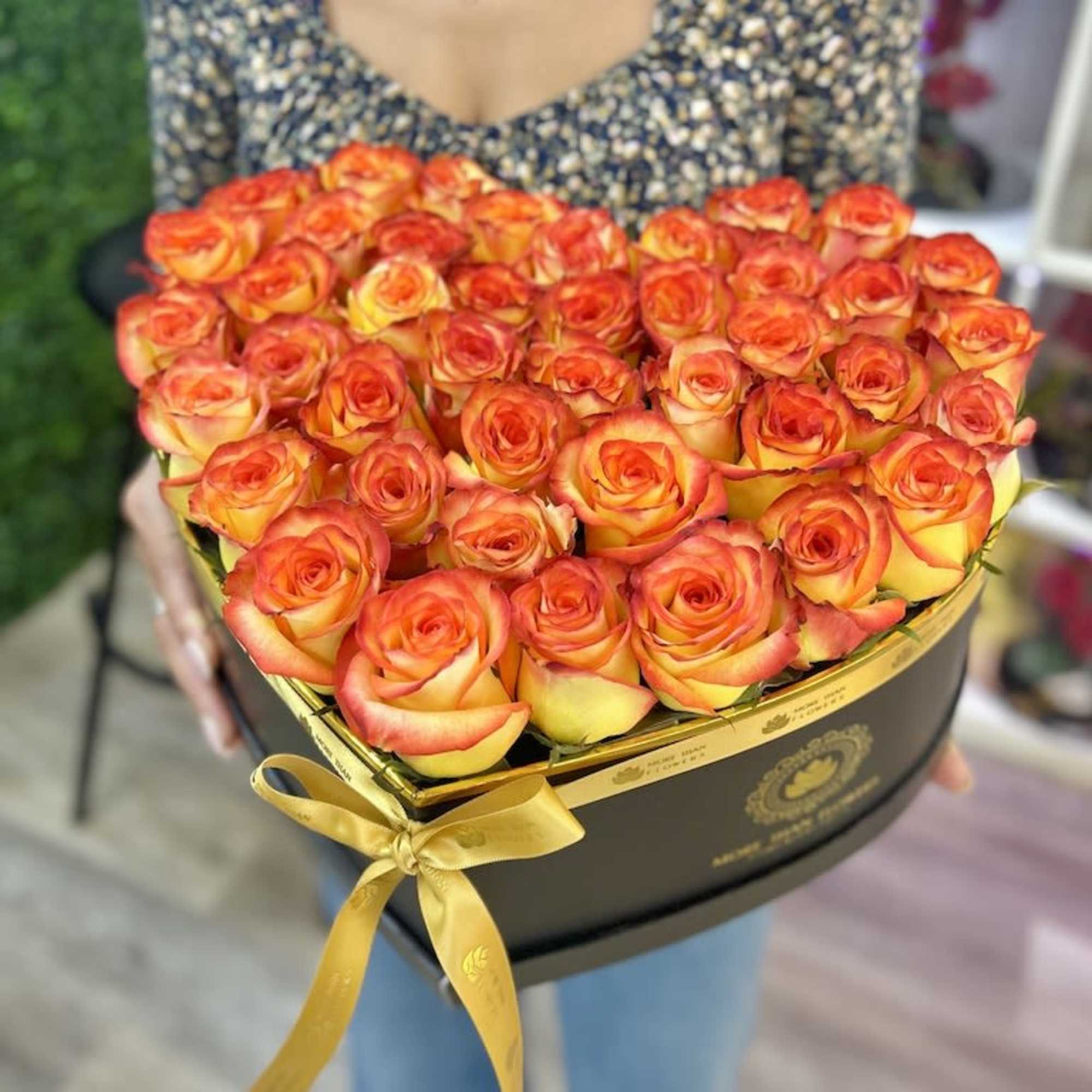 Heart-shaped black box filled with orange and yellow roses and tied with a gold ribbon