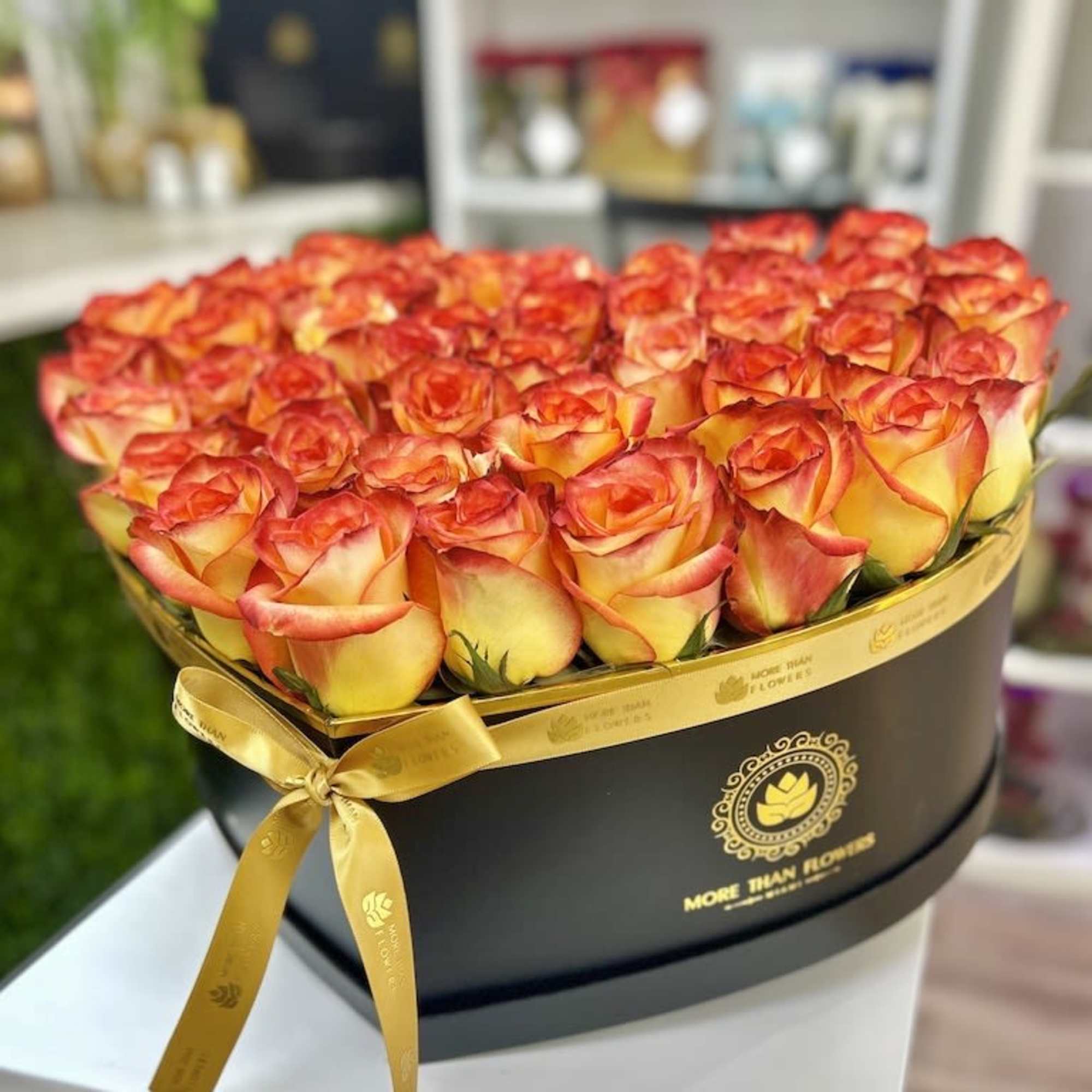 Heart-shaped black box filled with orange and yellow roses and tied with a gold ribbon