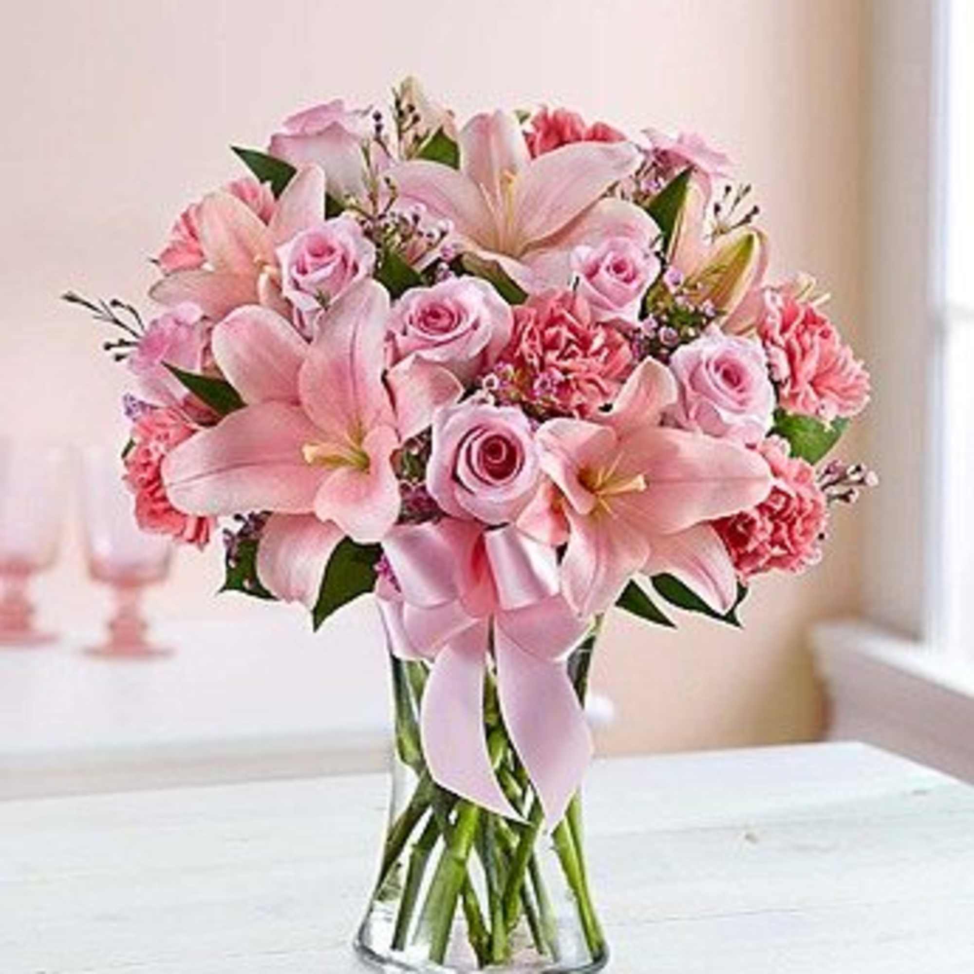 Pink lilies, roses, and carnations in a clear glass vase with a ribbon