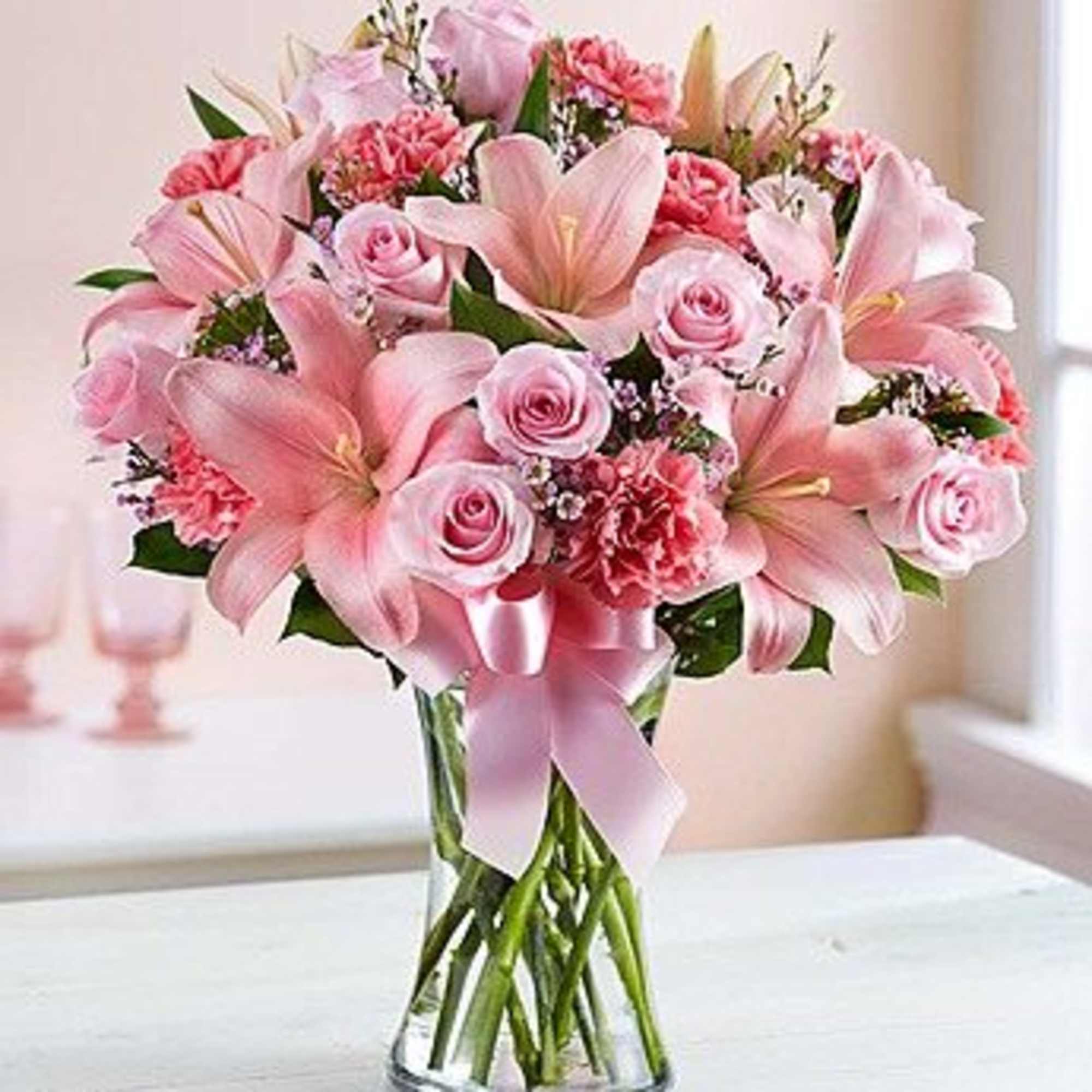 Pink lilies and roses arranged in a clear glass vase with a pink ribbon