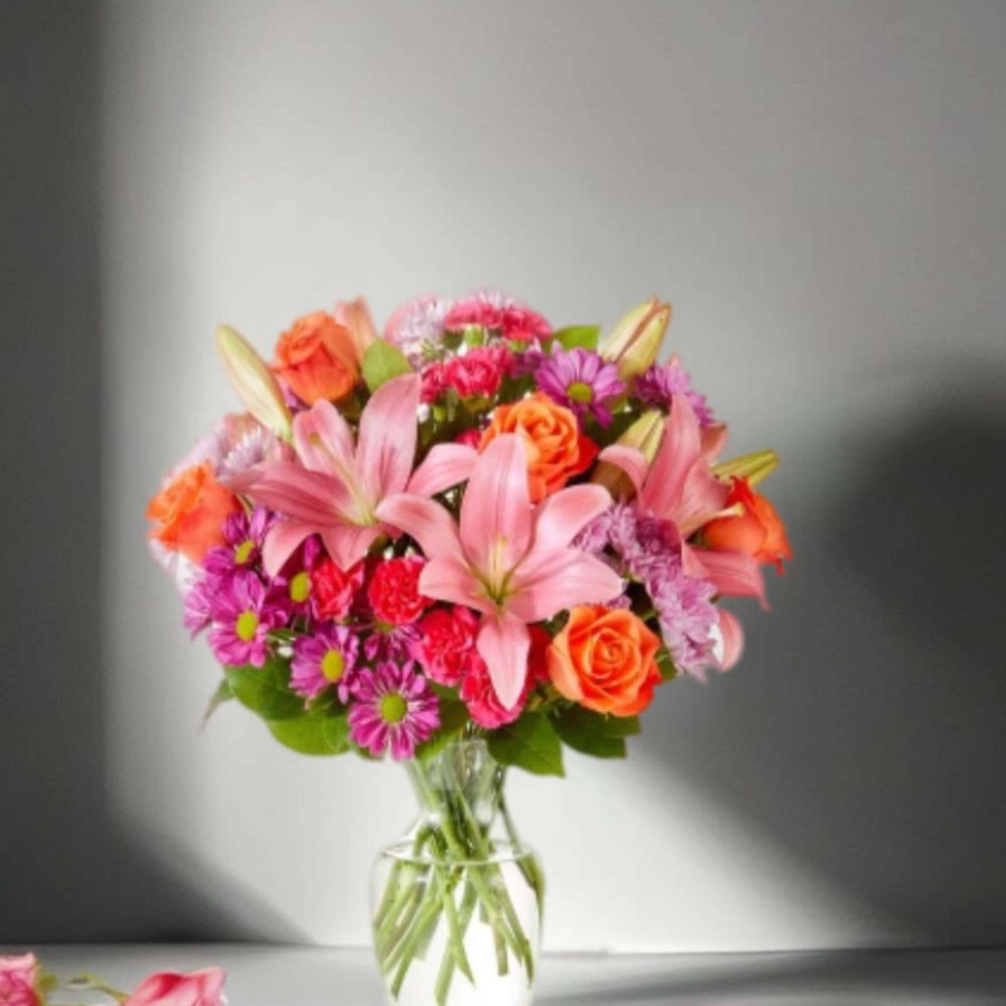 Mixed bouquet of pink lilies, orange roses, and purple daisies in a glass vase