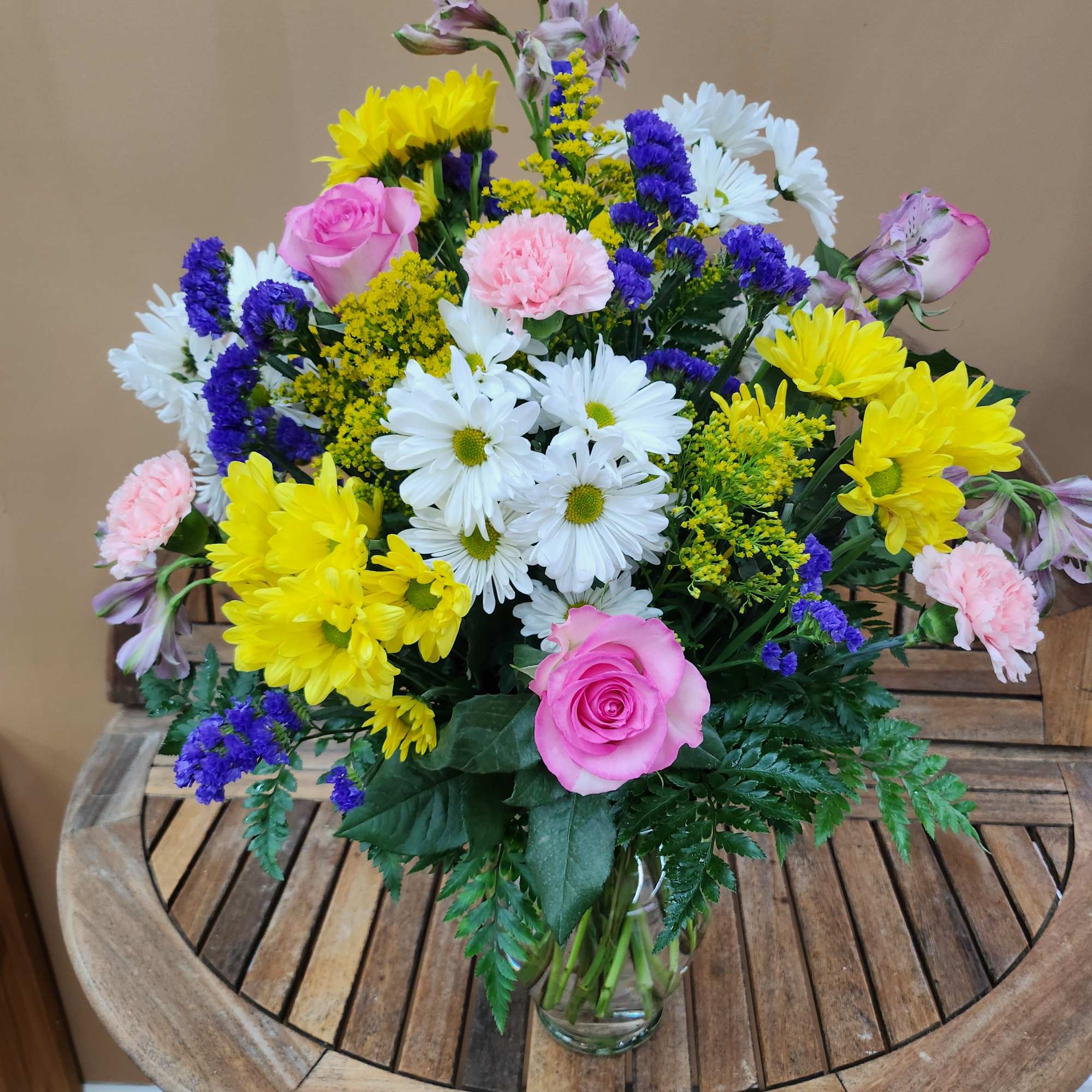 Mixed bouquet of pink roses, white daisies, yellow chrysanthemums, and purple filler flowers in a glass vase