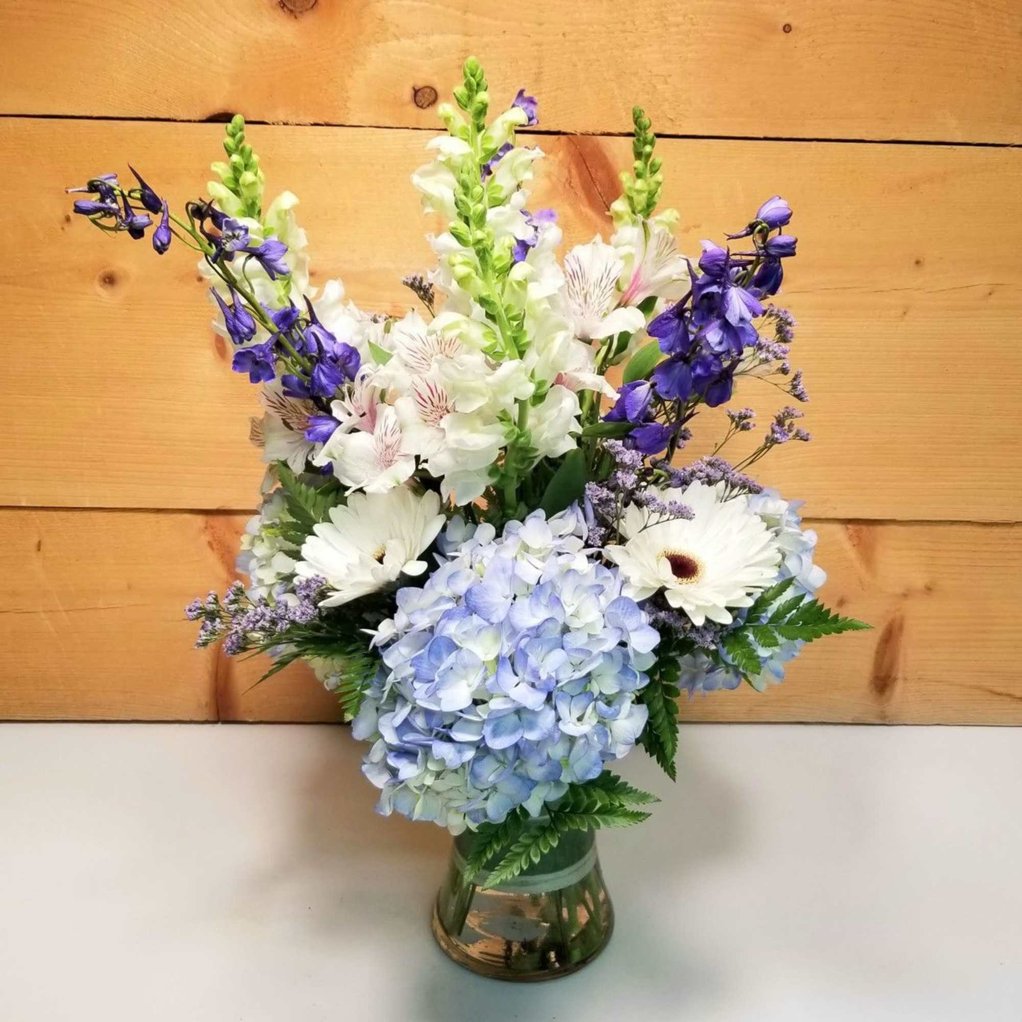 Mixed arrangement of white and purple flowers with blue hydrangeas in a clear glass vase