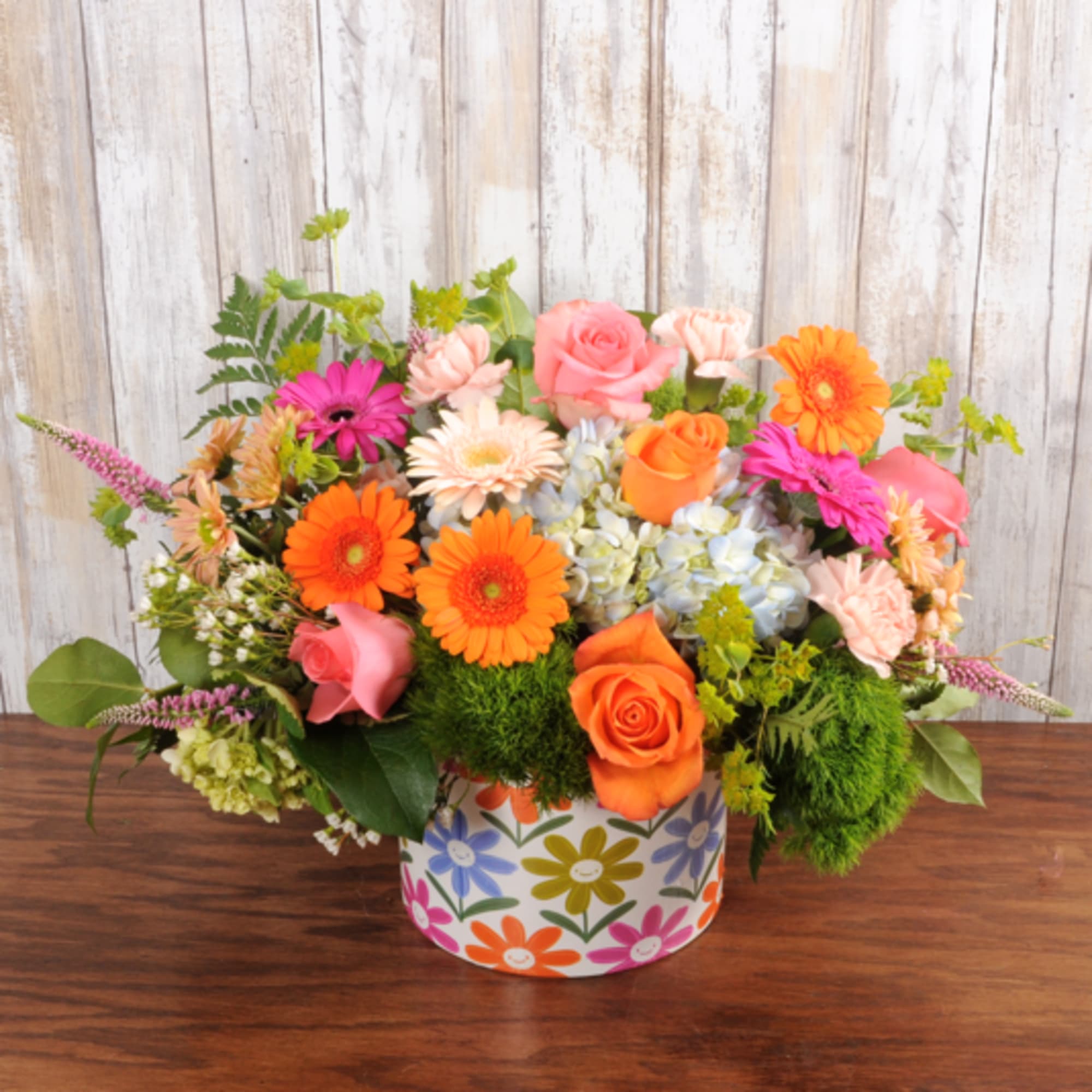 Low arrangement of orange gerbera daisies, pink roses, and mixed blooms in a colorful flower-print box.