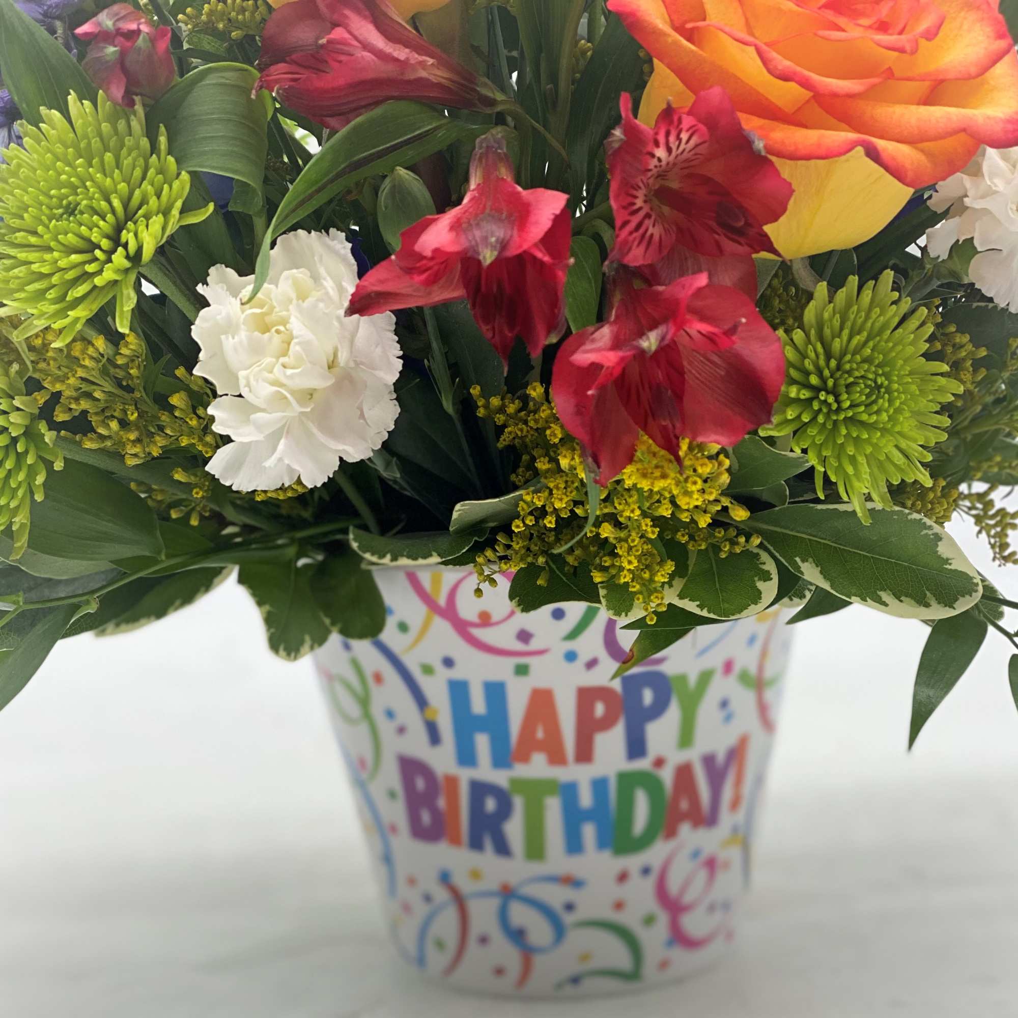 Bright mixed birthday flower arrangement with roses, carnations, and green mums in a Happy Birthday container