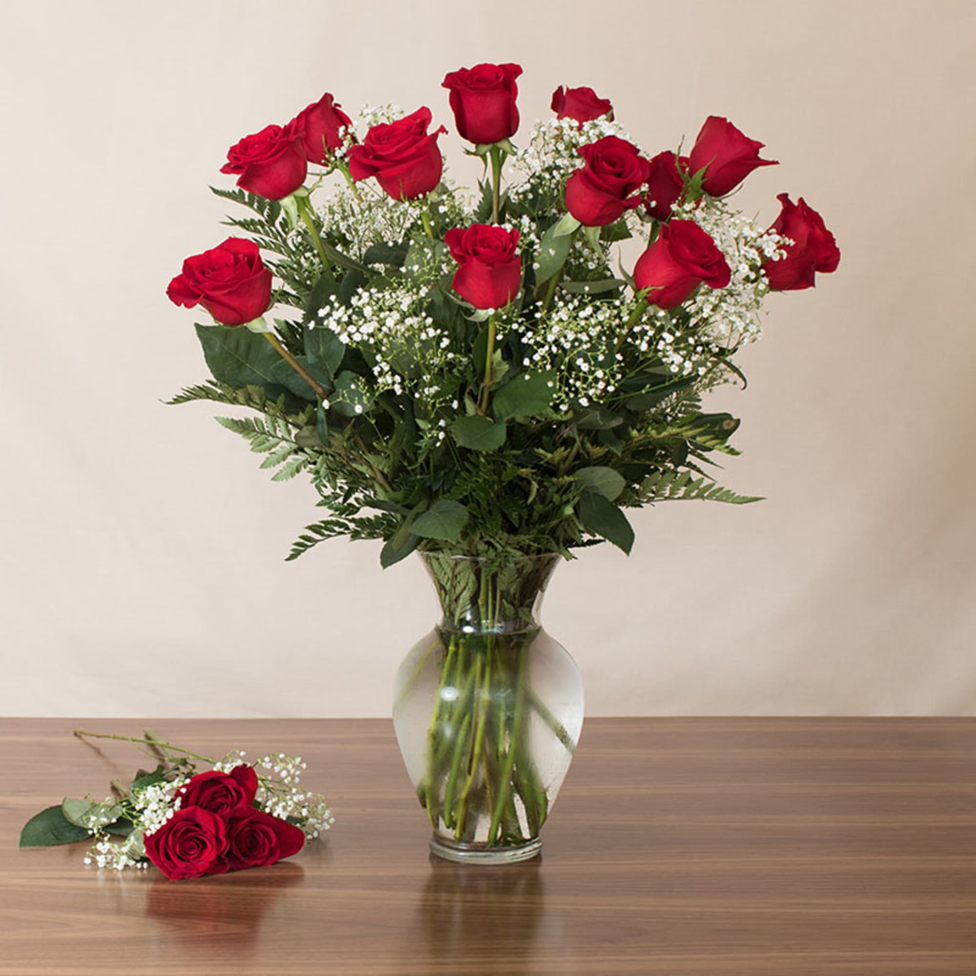 Tall arrangement of red roses and white filler flowers in a clear glass vase, with three roses laid beside it.