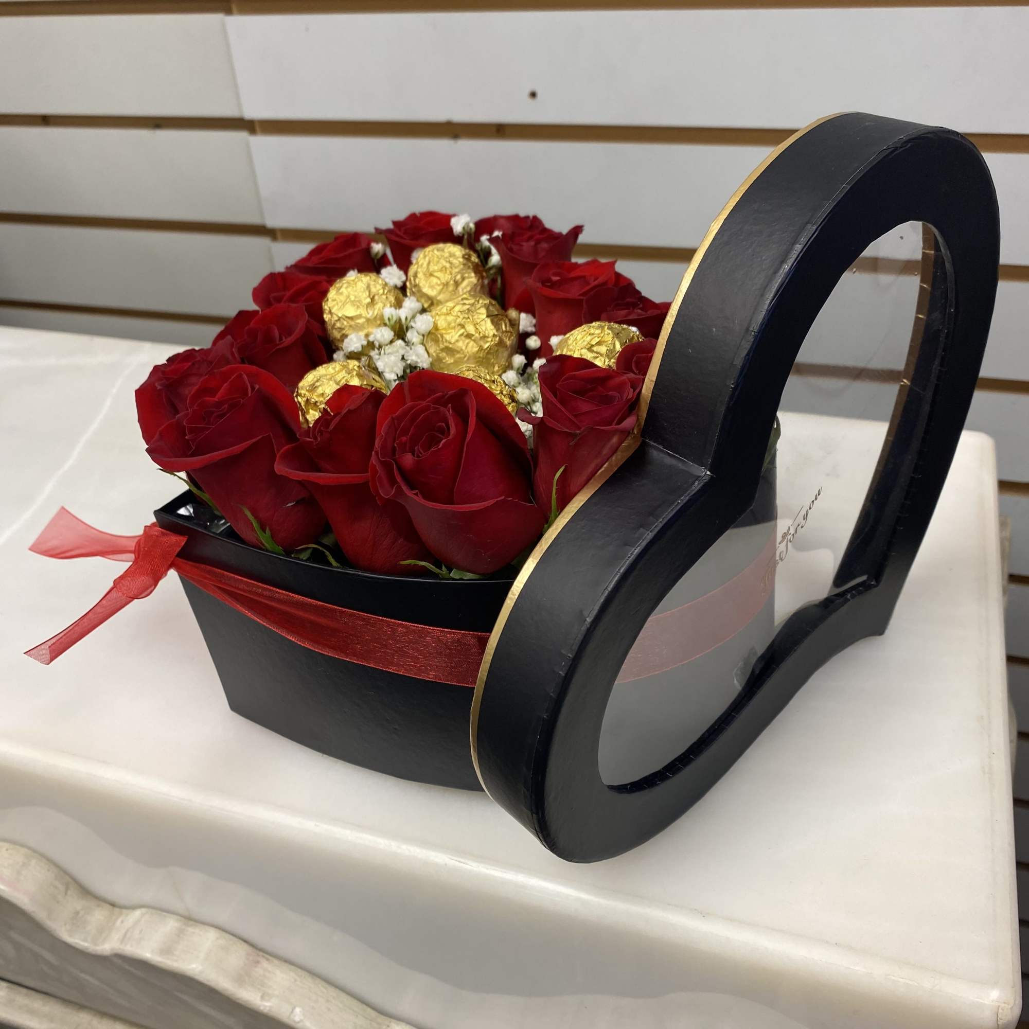 Heart-shaped black box filled with red roses, white filler flowers, and gold-wrapped chocolates.