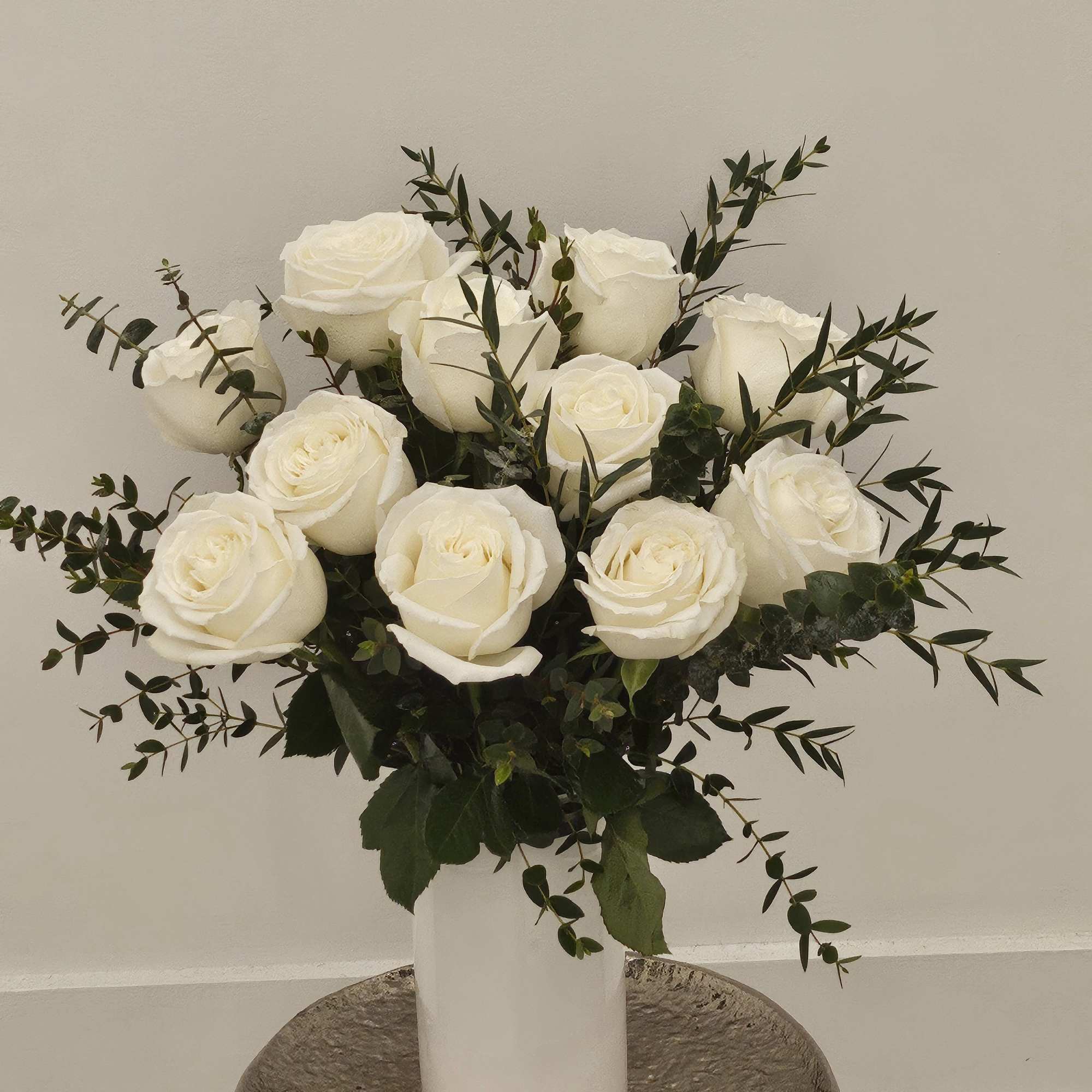 White roses arranged in a tall white vase with dark greenery