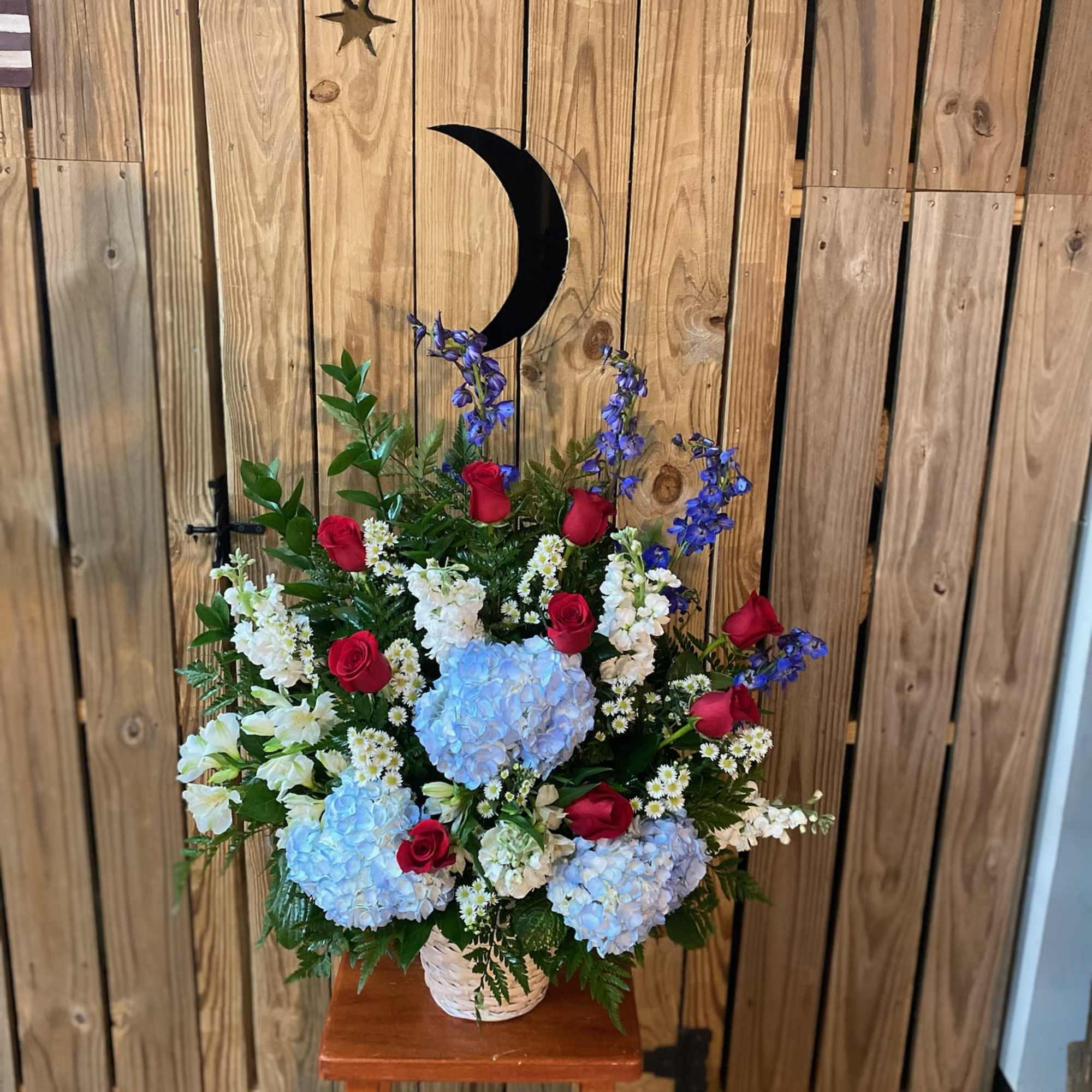 A red, white, and blue tribute funeral basket featuring red roses, blue