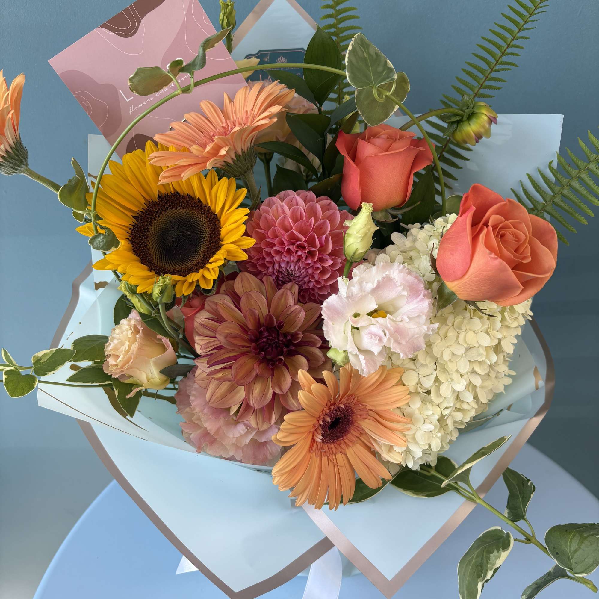 Mixed bouquet with sunflowers, roses, and dahlias wrapped in pale blue paper