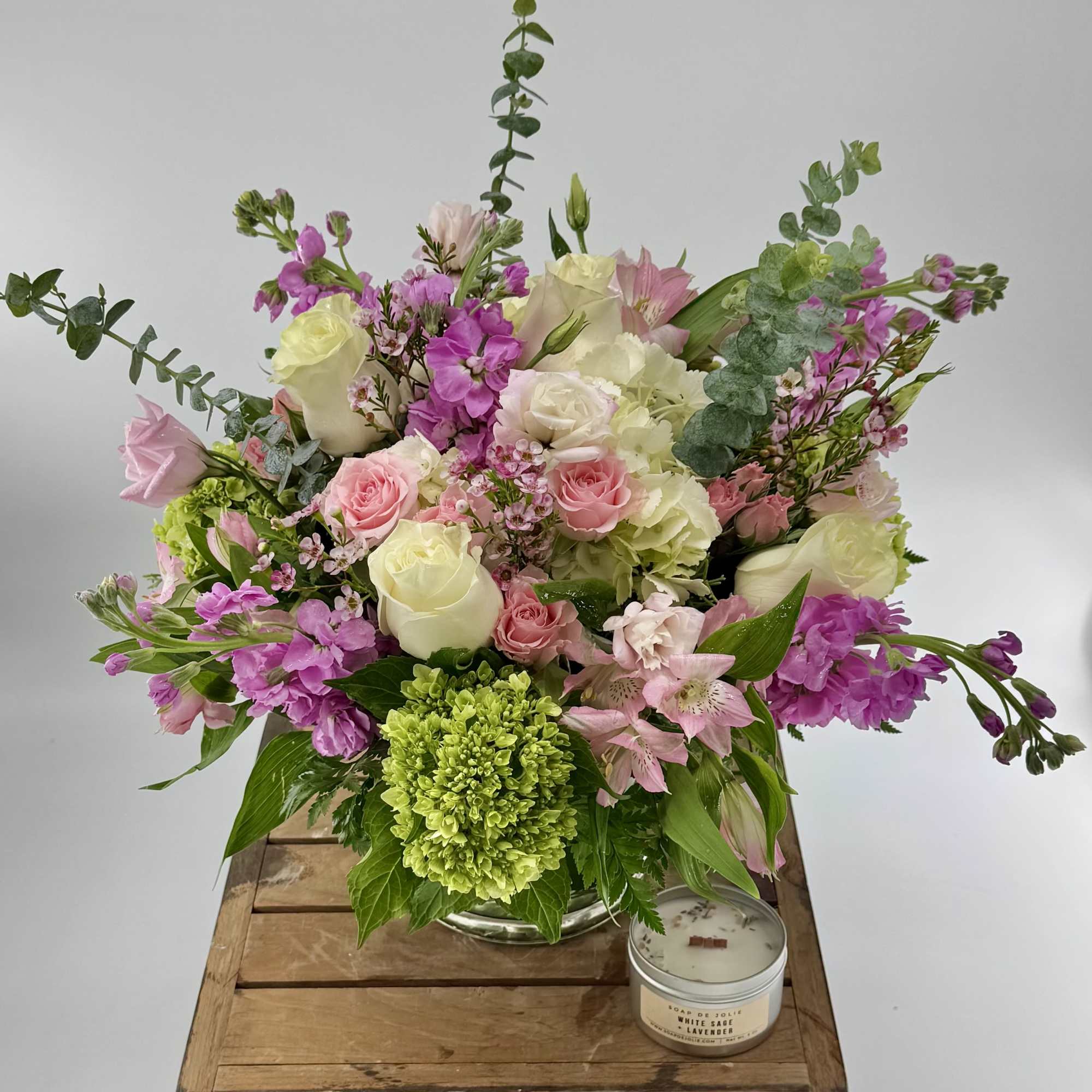 Pink and white mixed flower arrangement with roses and stock in a clear vase beside a small candle.