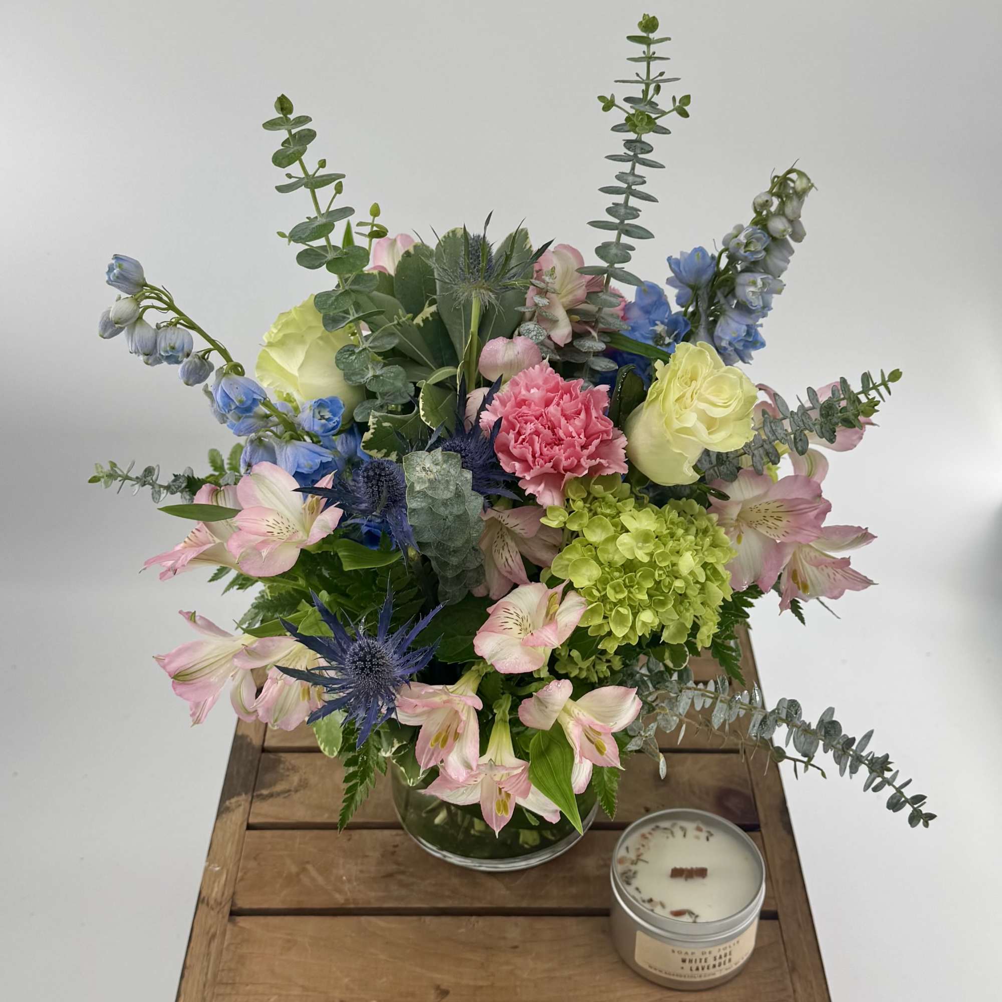 Mixed bouquet of pink, blue, and green flowers in a clear vase with a small candle beside it.