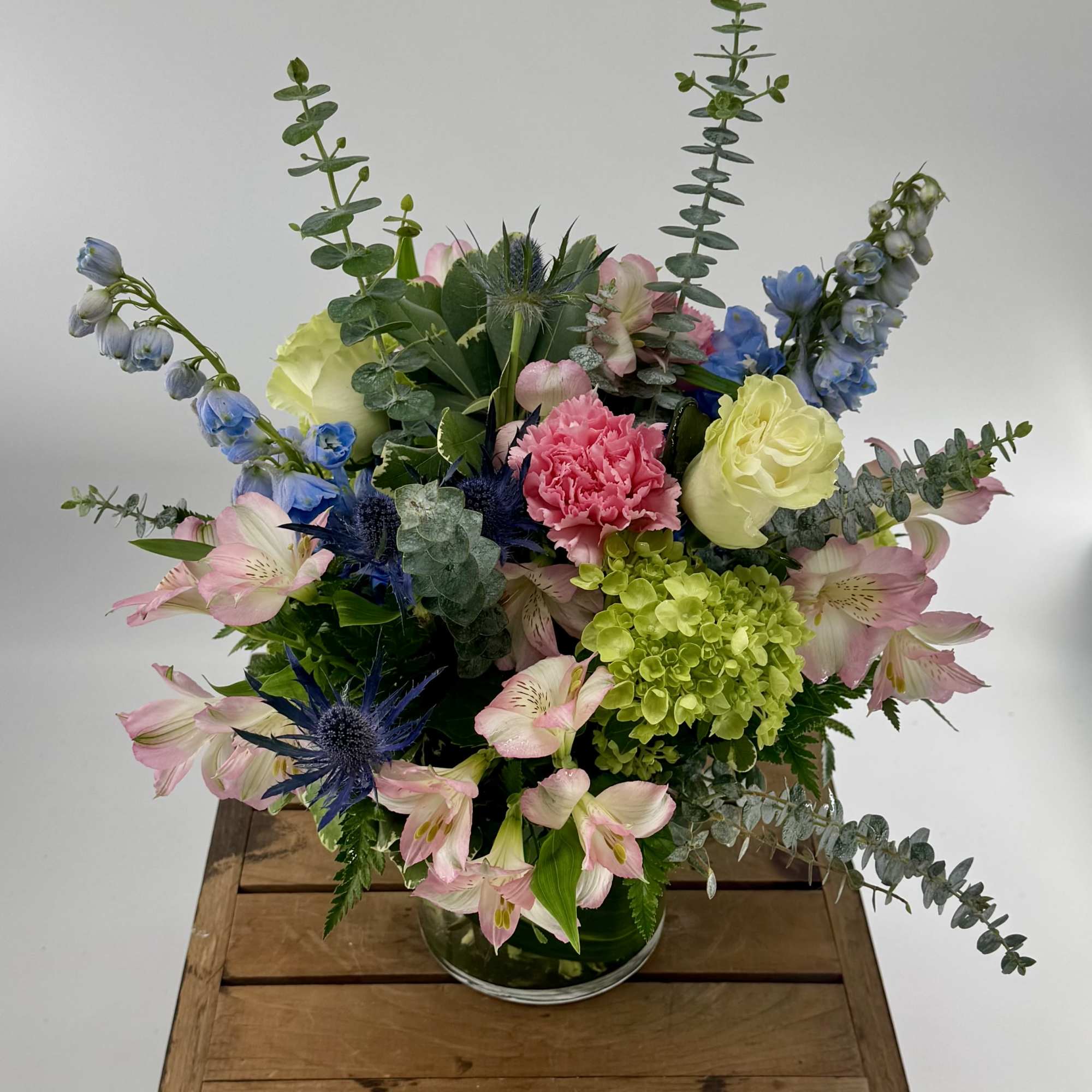 Mixed arrangement of pink, blue, green, and cream flowers in a clear glass vase on a wooden crate.