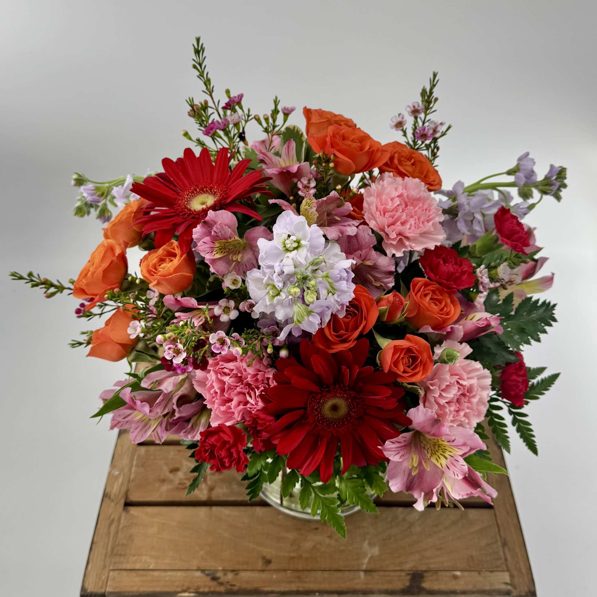 Bright mixed bouquet of red, pink, and orange flowers in a glass vase on a wooden crate