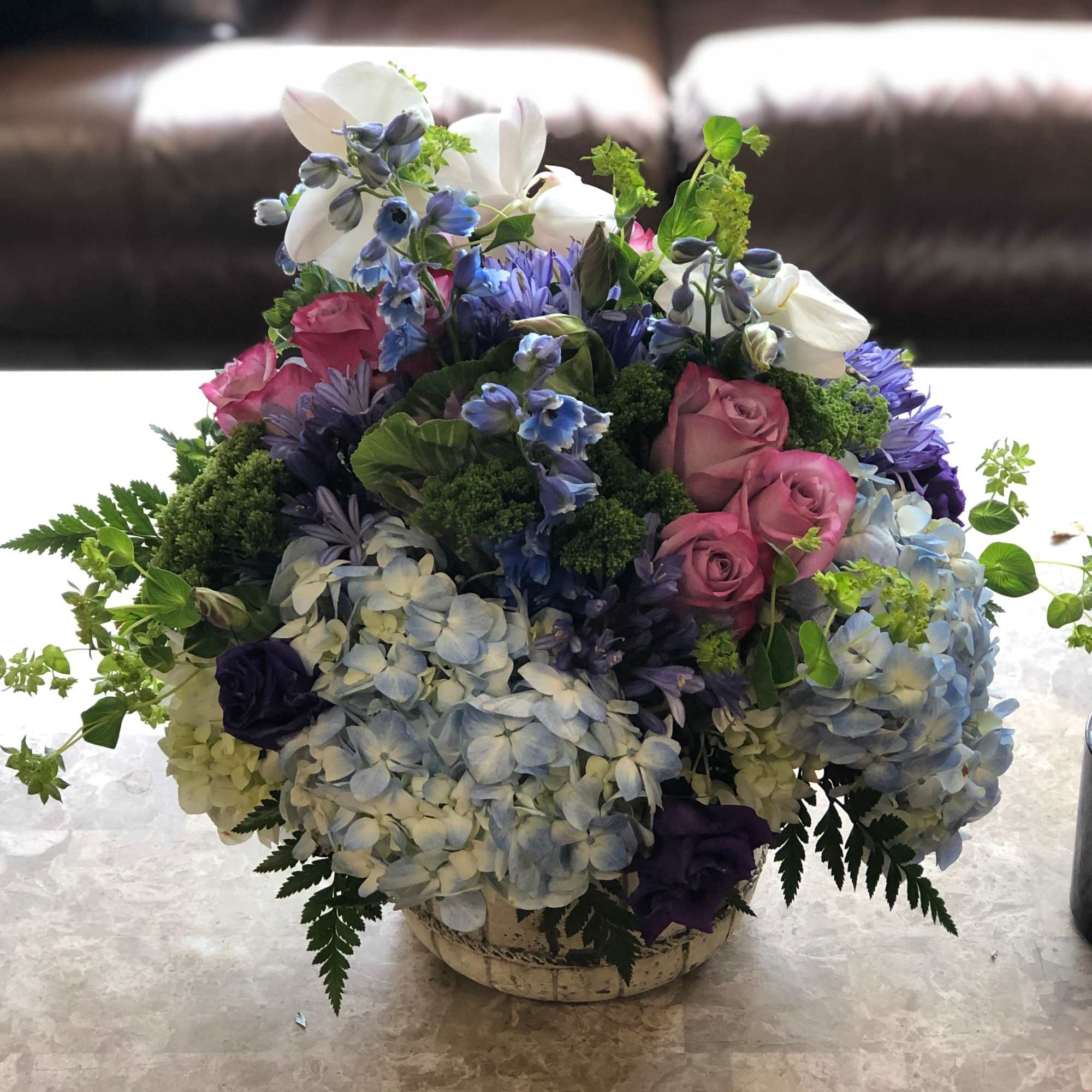 Round arrangement of blue hydrangeas, pink roses, purple blooms, and white orchids in a ceramic bowl