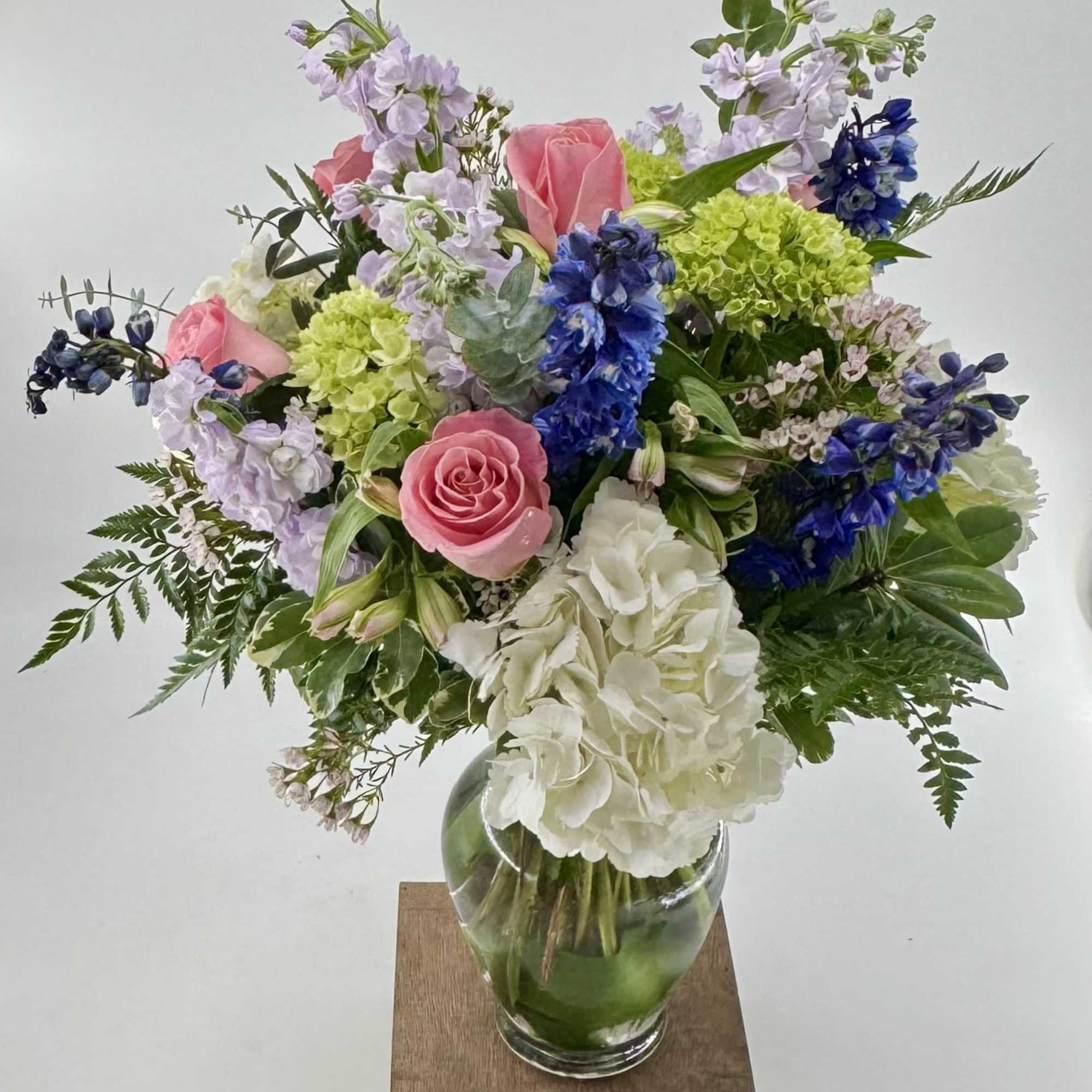 Mixed bouquet of pink roses, blue delphinium, and white hydrangeas arranged in a clear glass vase.