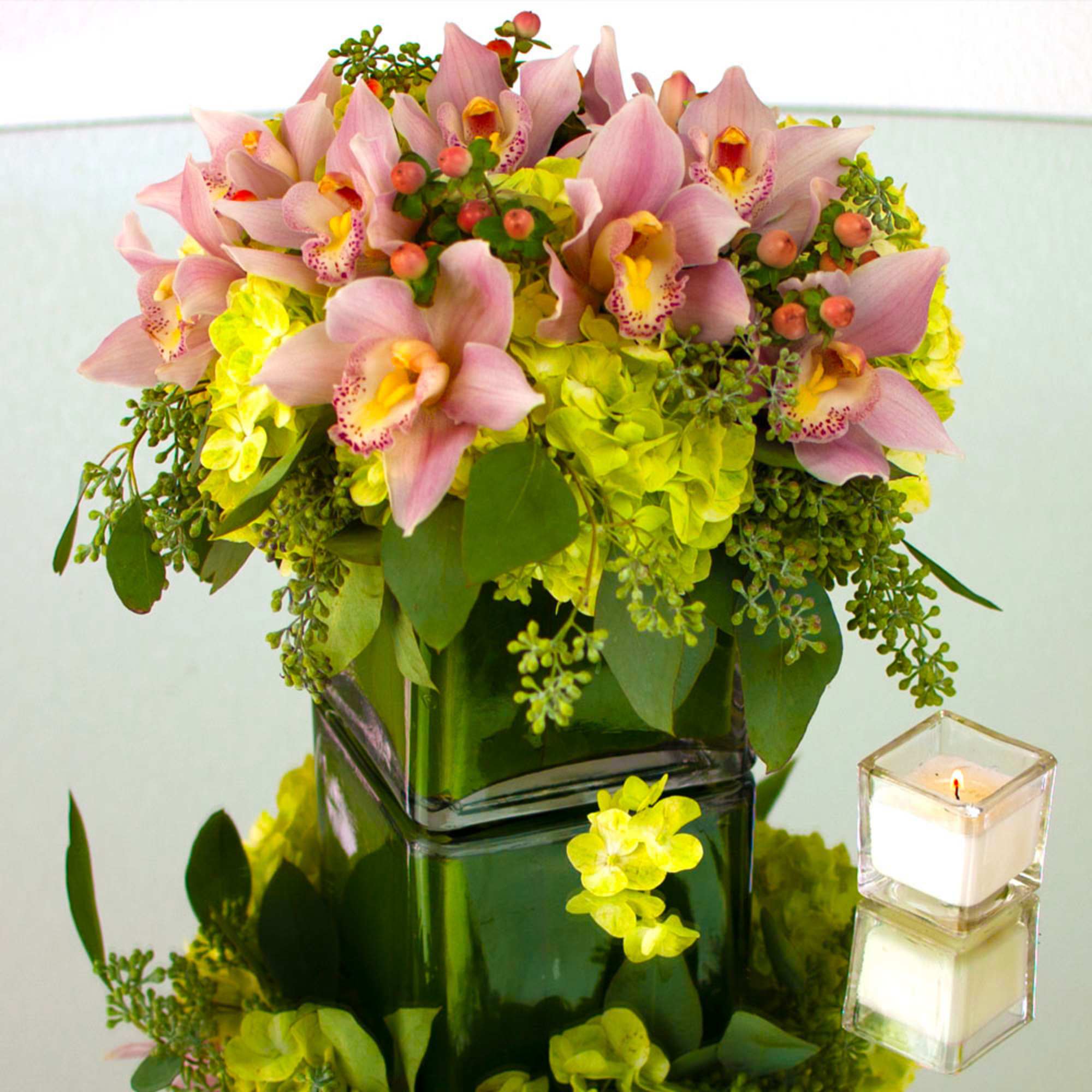 Pink cymbidium orchids and yellow-green hydrangeas in a green glass cube vase beside a lit votive candle