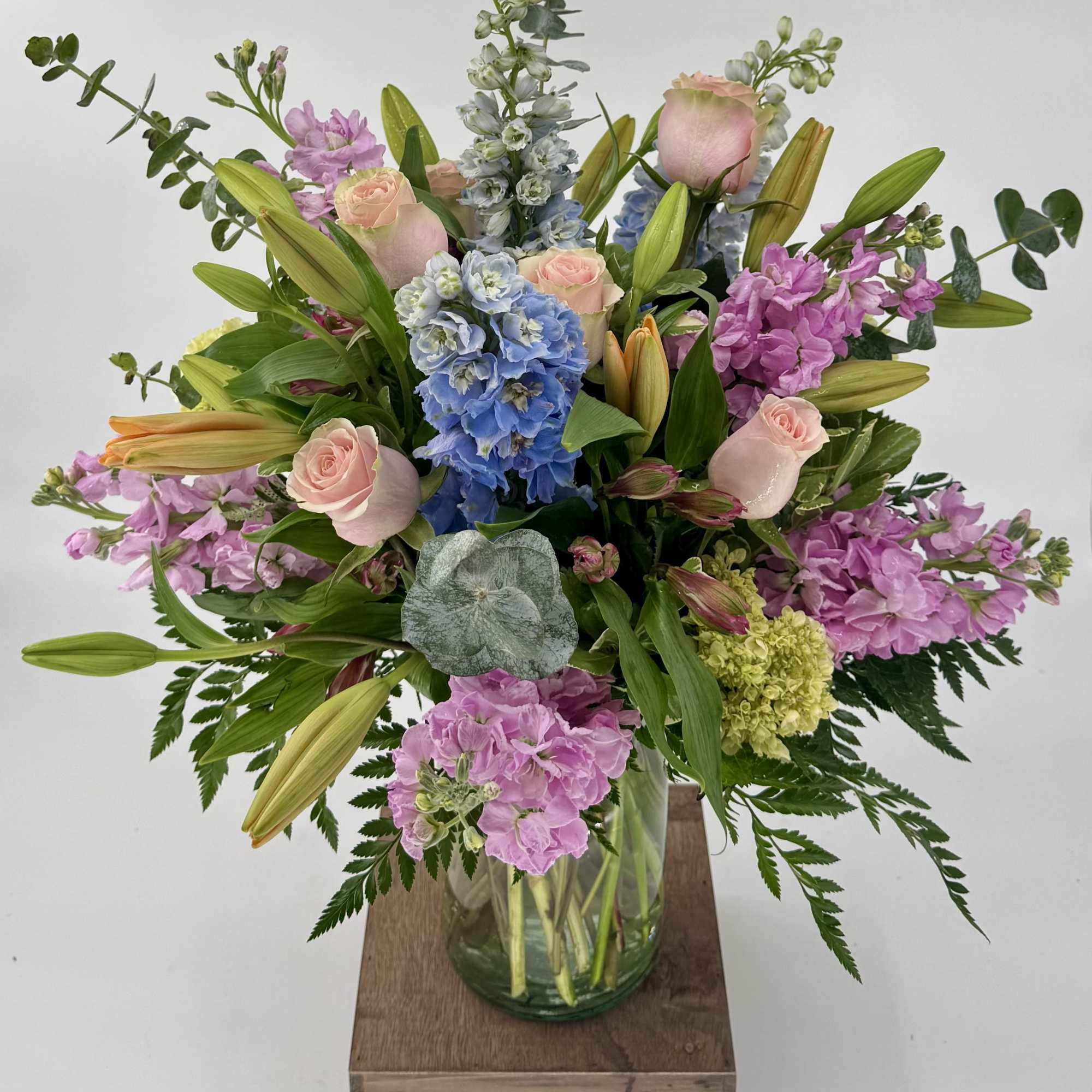 Tall pastel arrangement of pink roses, lilies, blue delphinium and stock in a clear glass vase