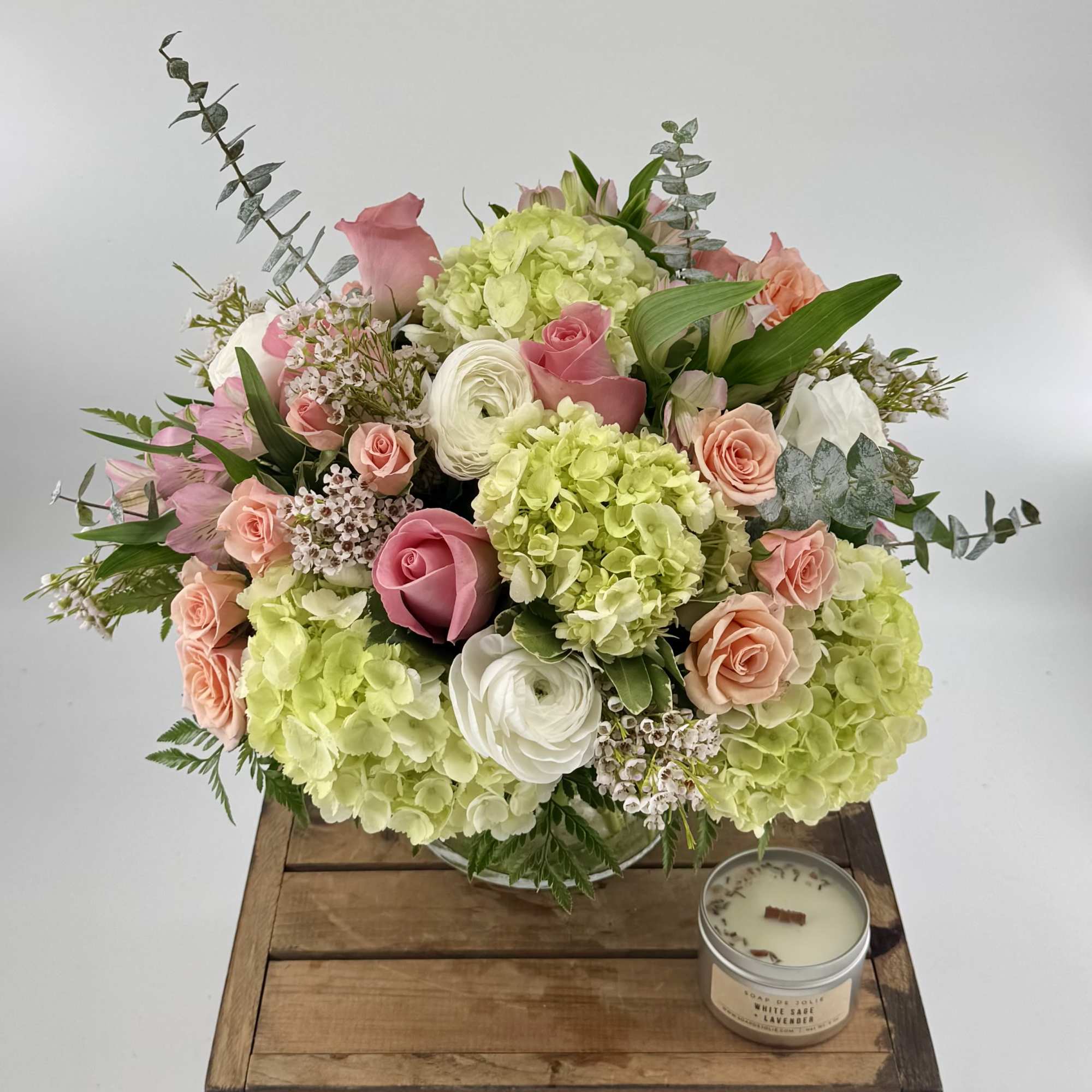 Pink and peach roses with green hydrangeas in a clear vase beside a small candle.