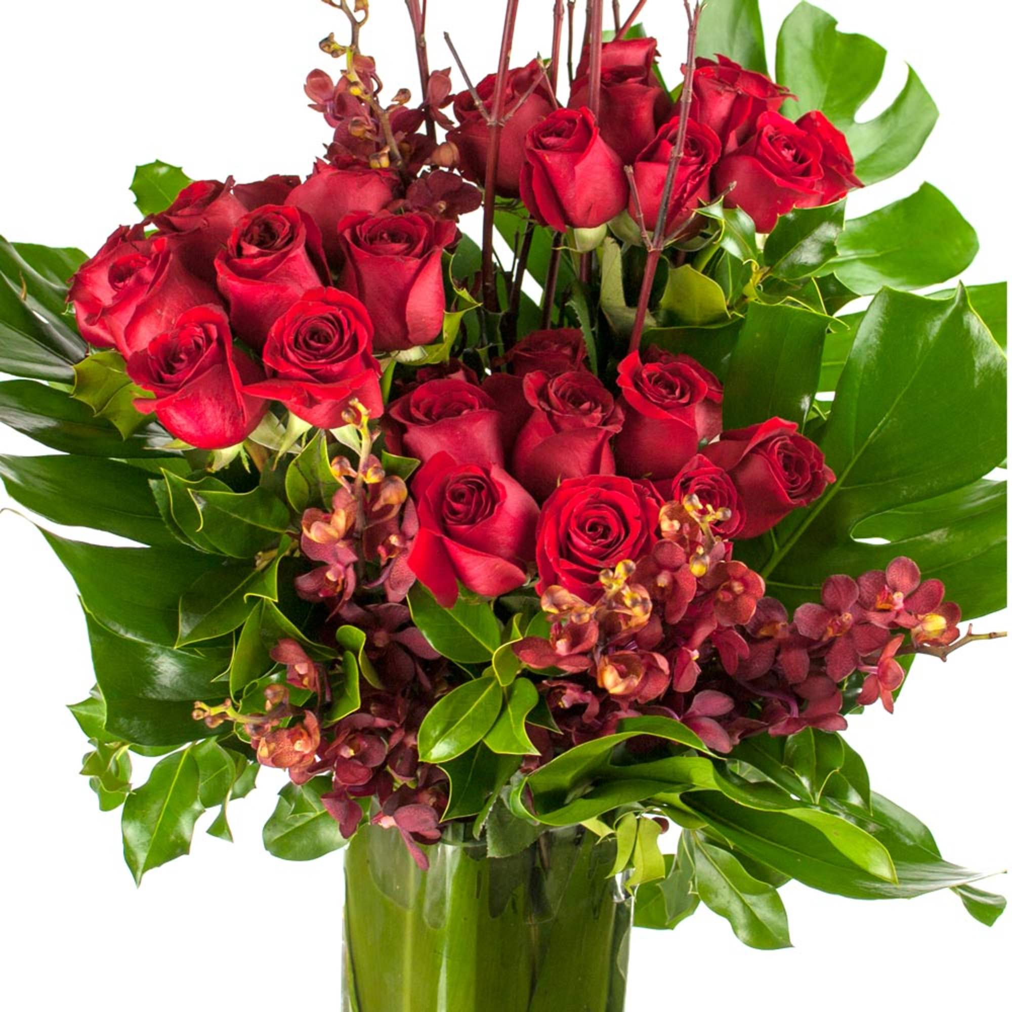 Tall arrangement of red roses and burgundy orchids in a clear glass vase with bold glossy foliage.