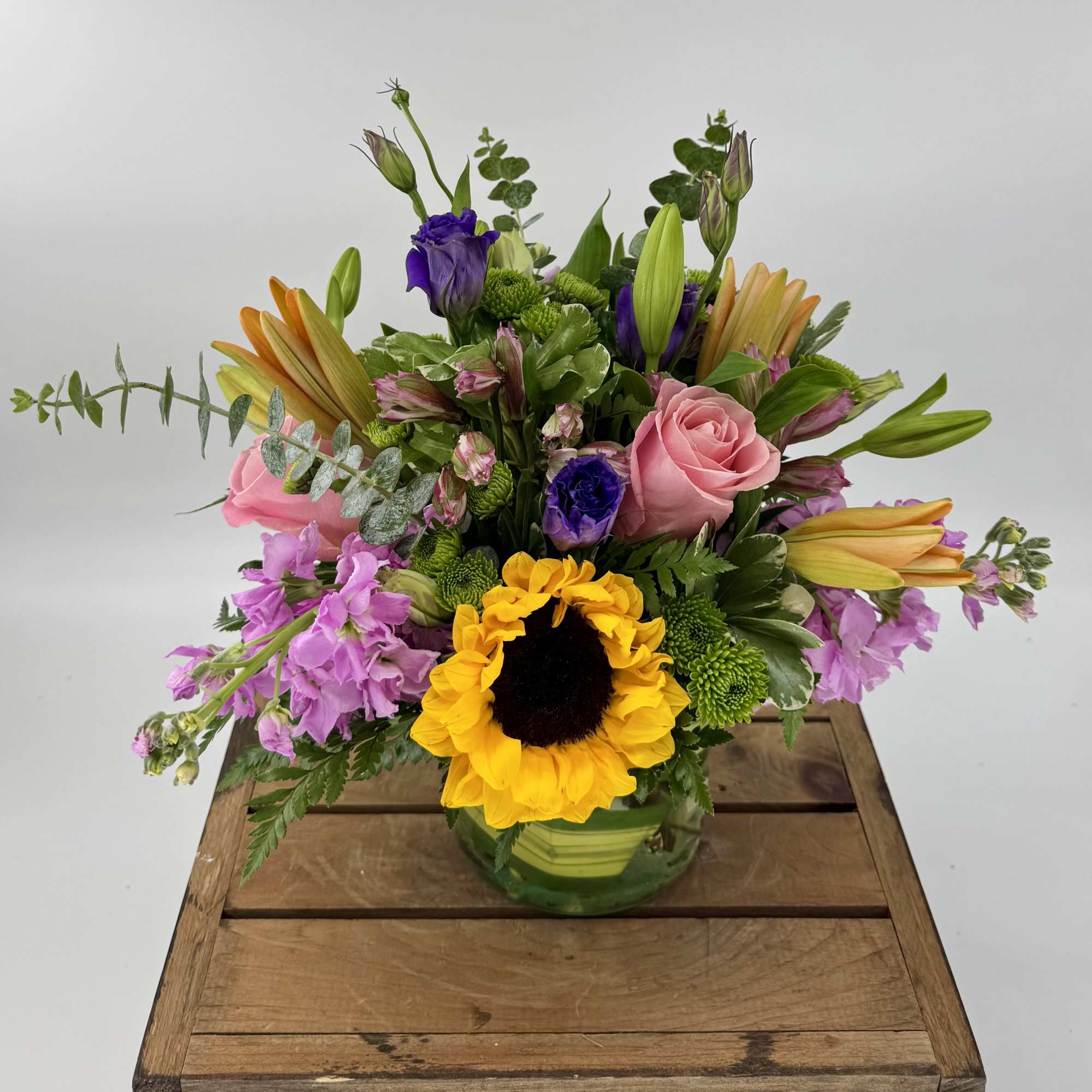 Round vase arrangement with yellow sunflower, pink roses, orange lilies, and purple blooms