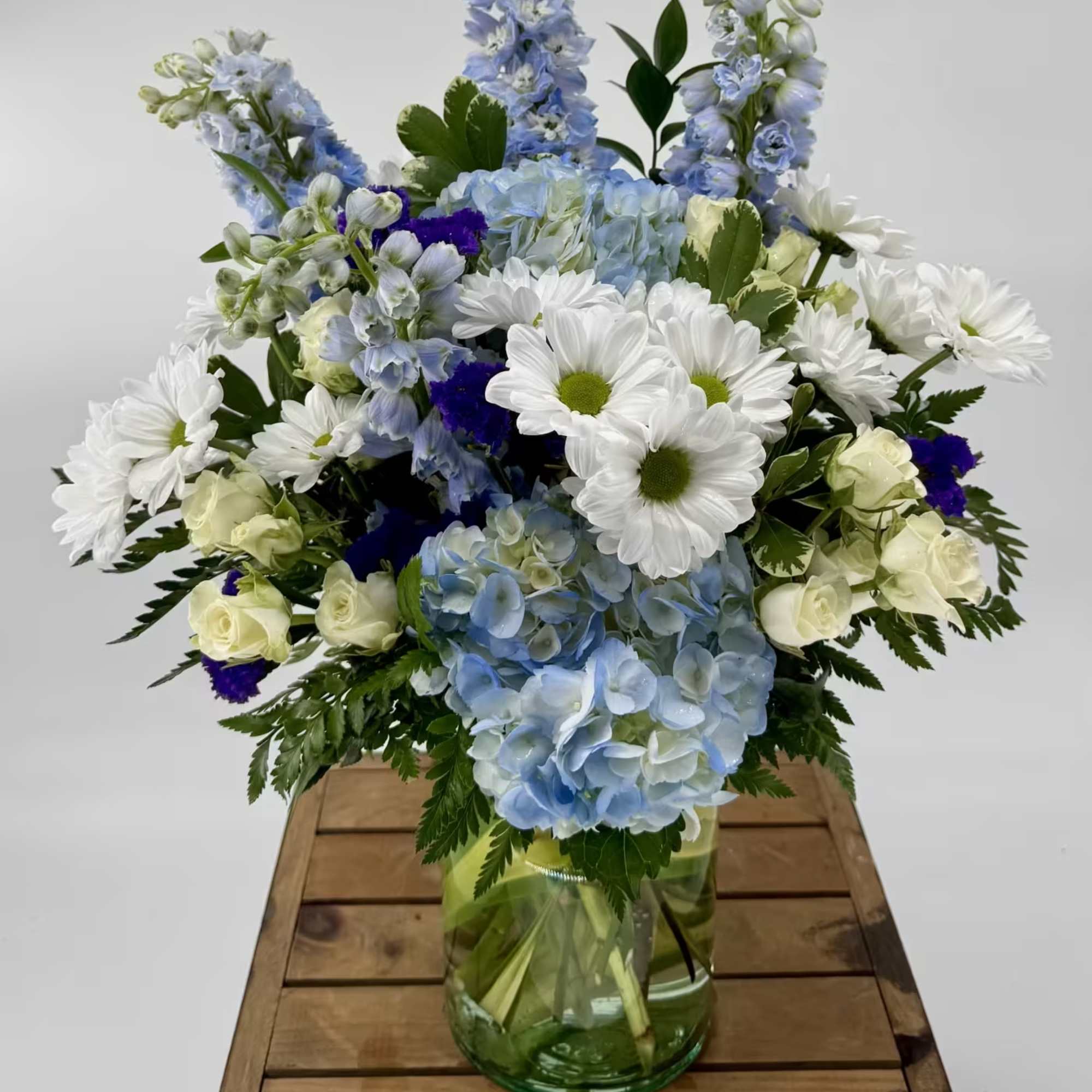 Blue hydrangeas, white daisies, and roses in a green glass vase on a wooden crate