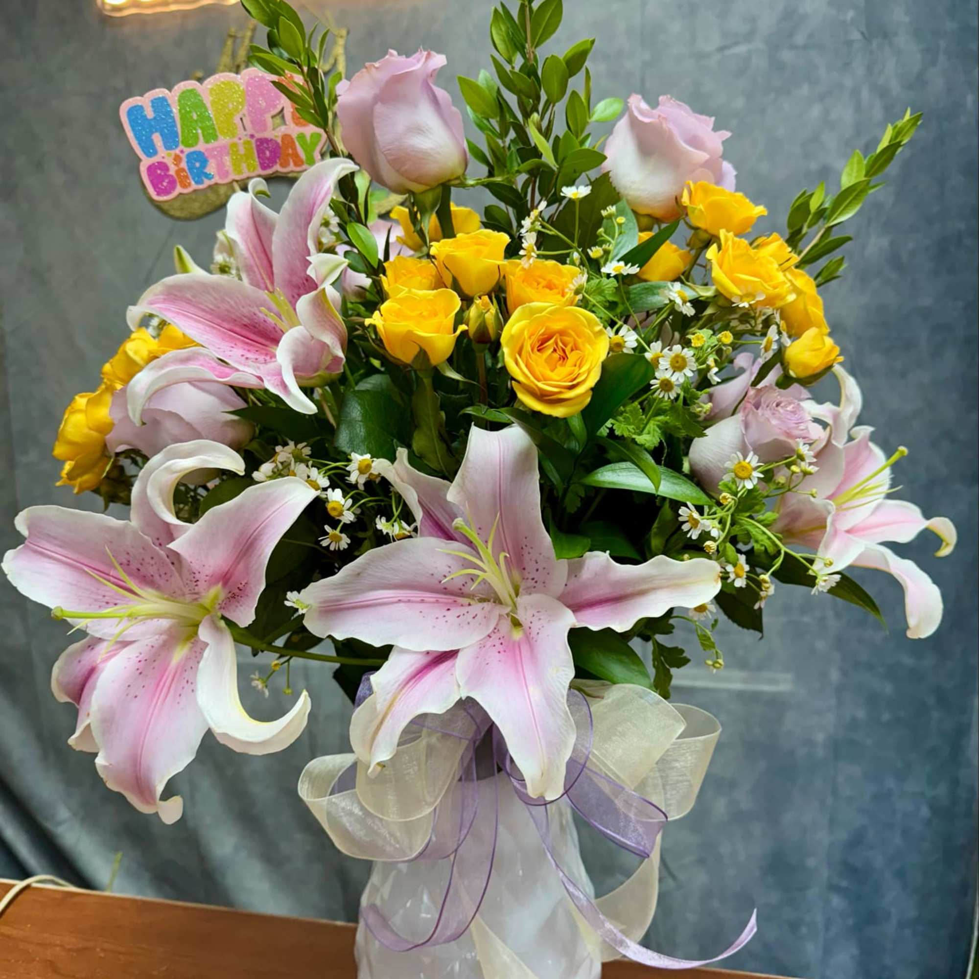 Pink lilies and yellow roses arranged in a white vase with a birthday sign