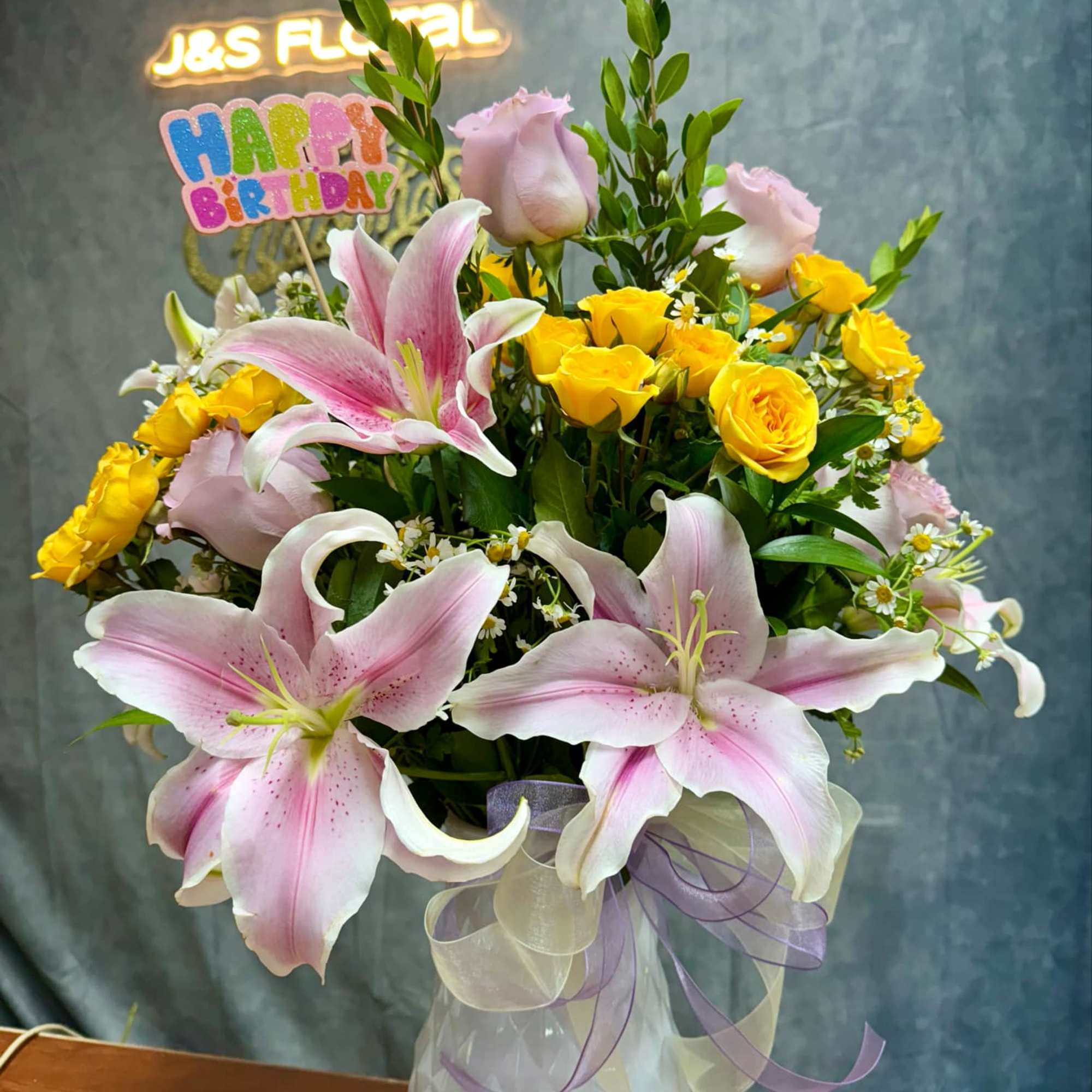 Pink lilies and yellow roses in a white vase with a birthday sign