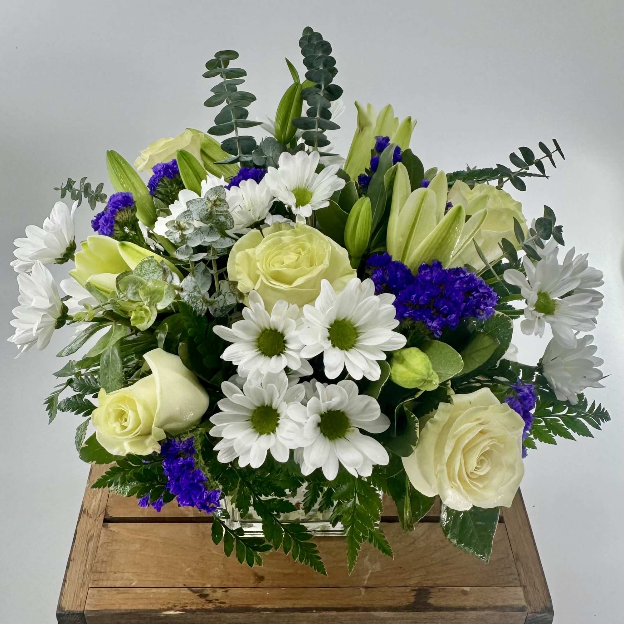 Mixed arrangement of white roses, daisies, and lilies with purple flowers in a square glass vase.