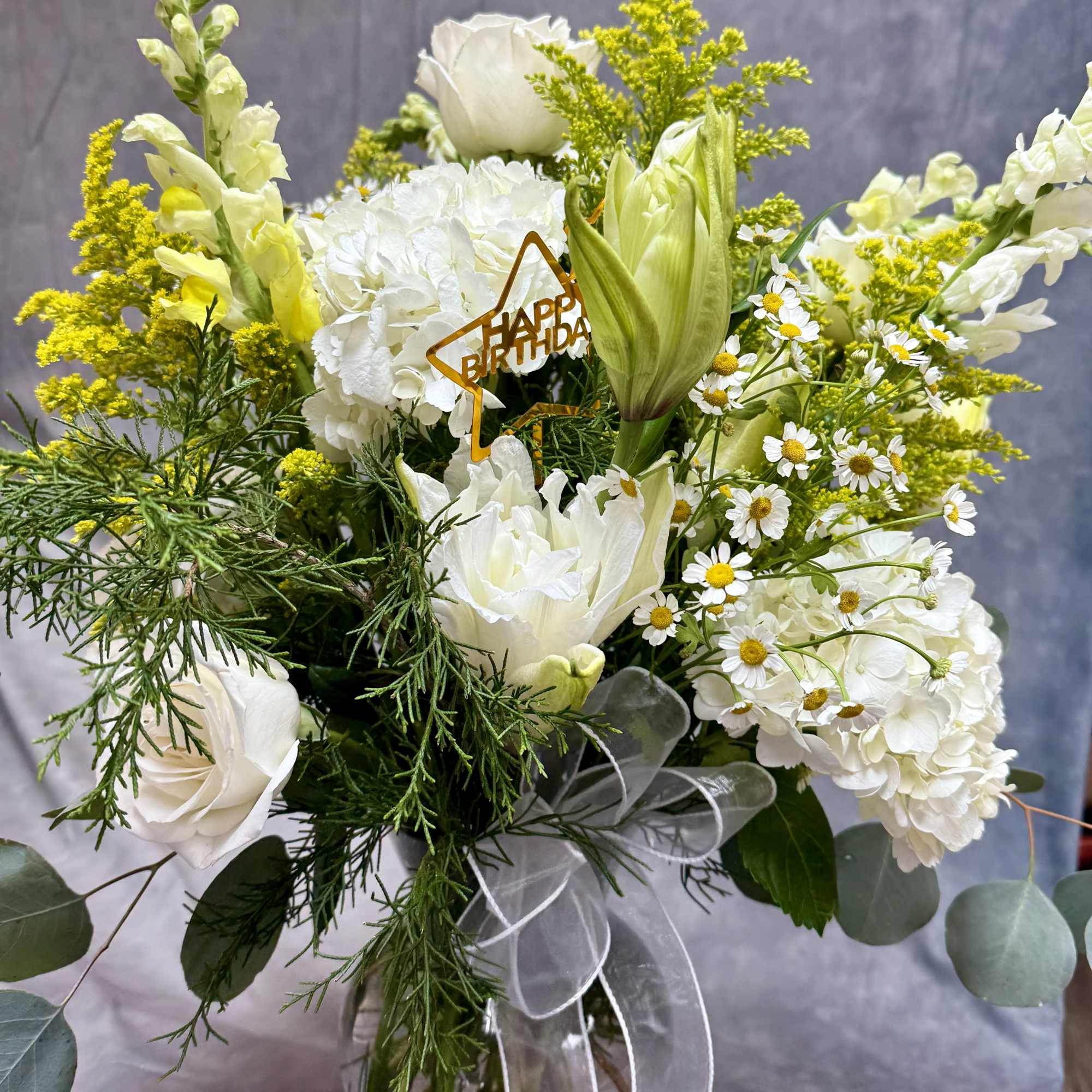 White floral bouquet with yellow accents and a birthday pick in a glass vase