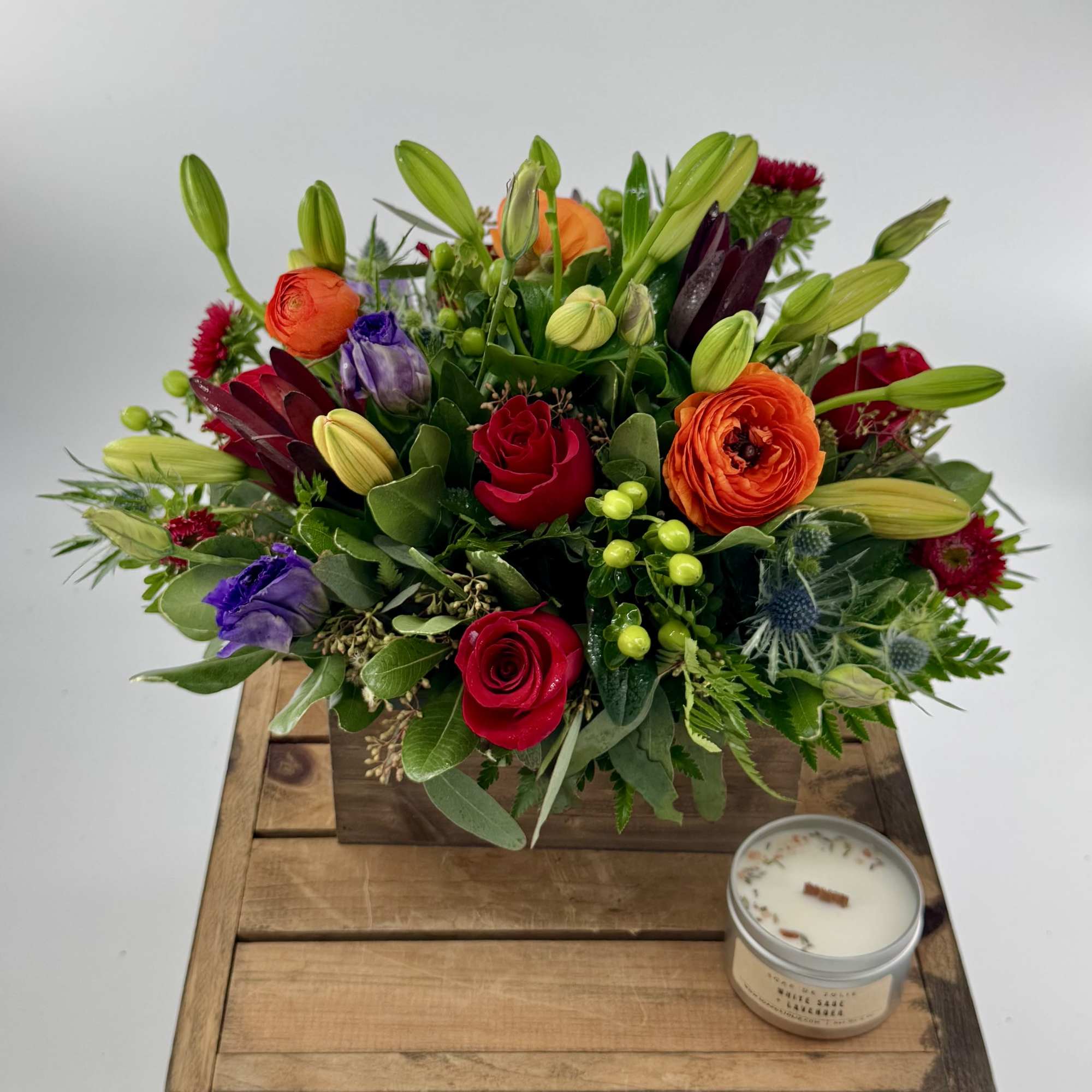 Mixed bouquet with roses, lilies, and purple blooms in a wooden box beside a candle