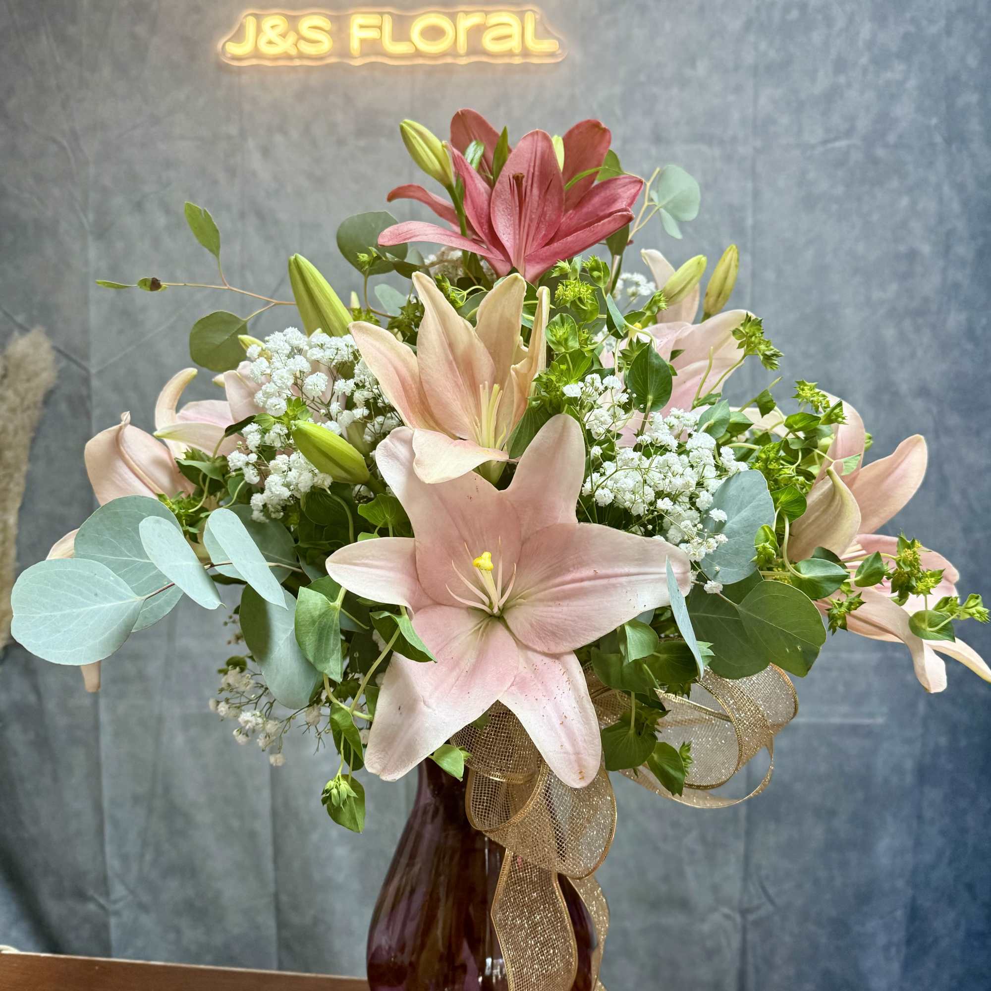 Pink lilies and baby's breath in a burgundy vase with a gold ribbon