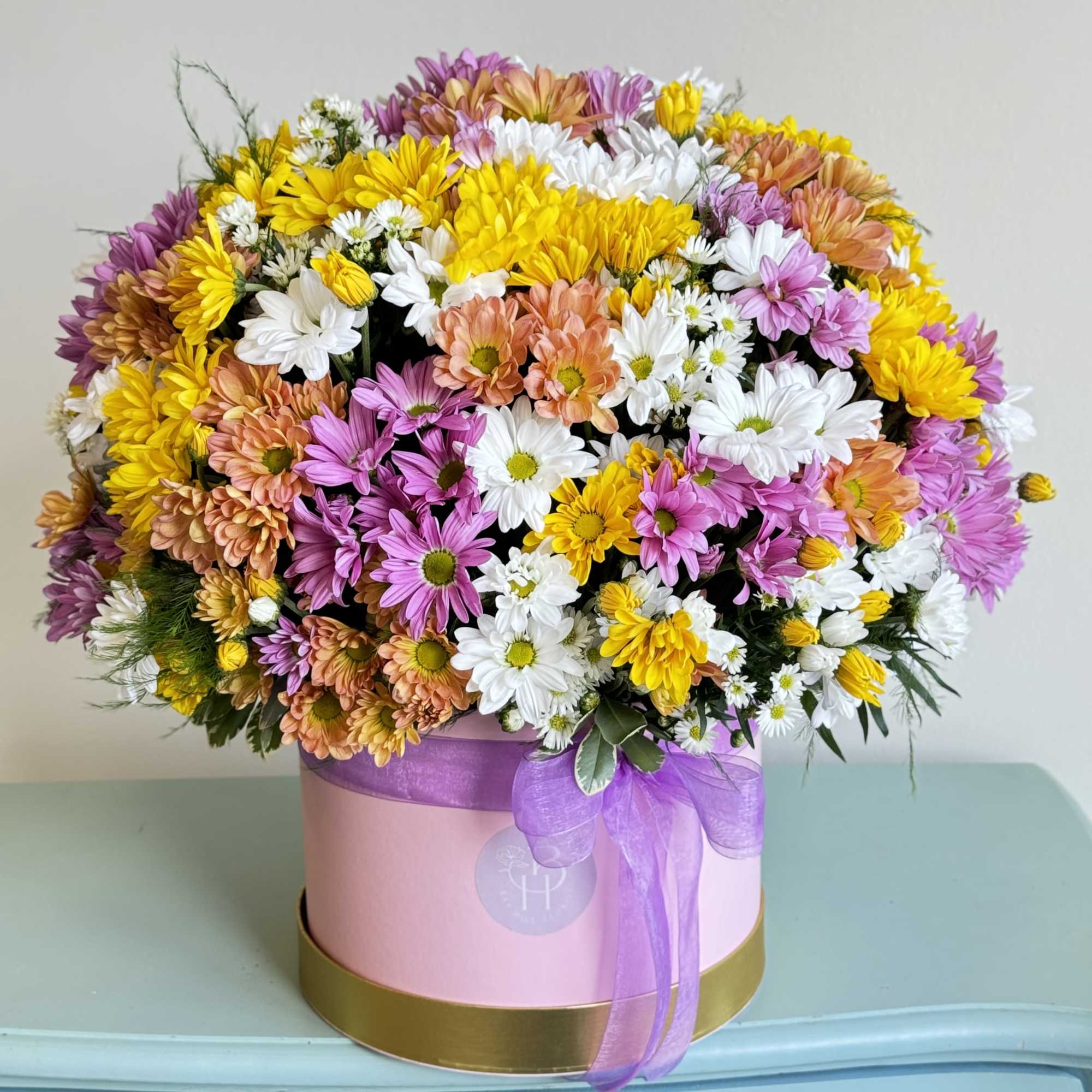 Mixed daisy bouquet in a pink round box with a purple ribbon