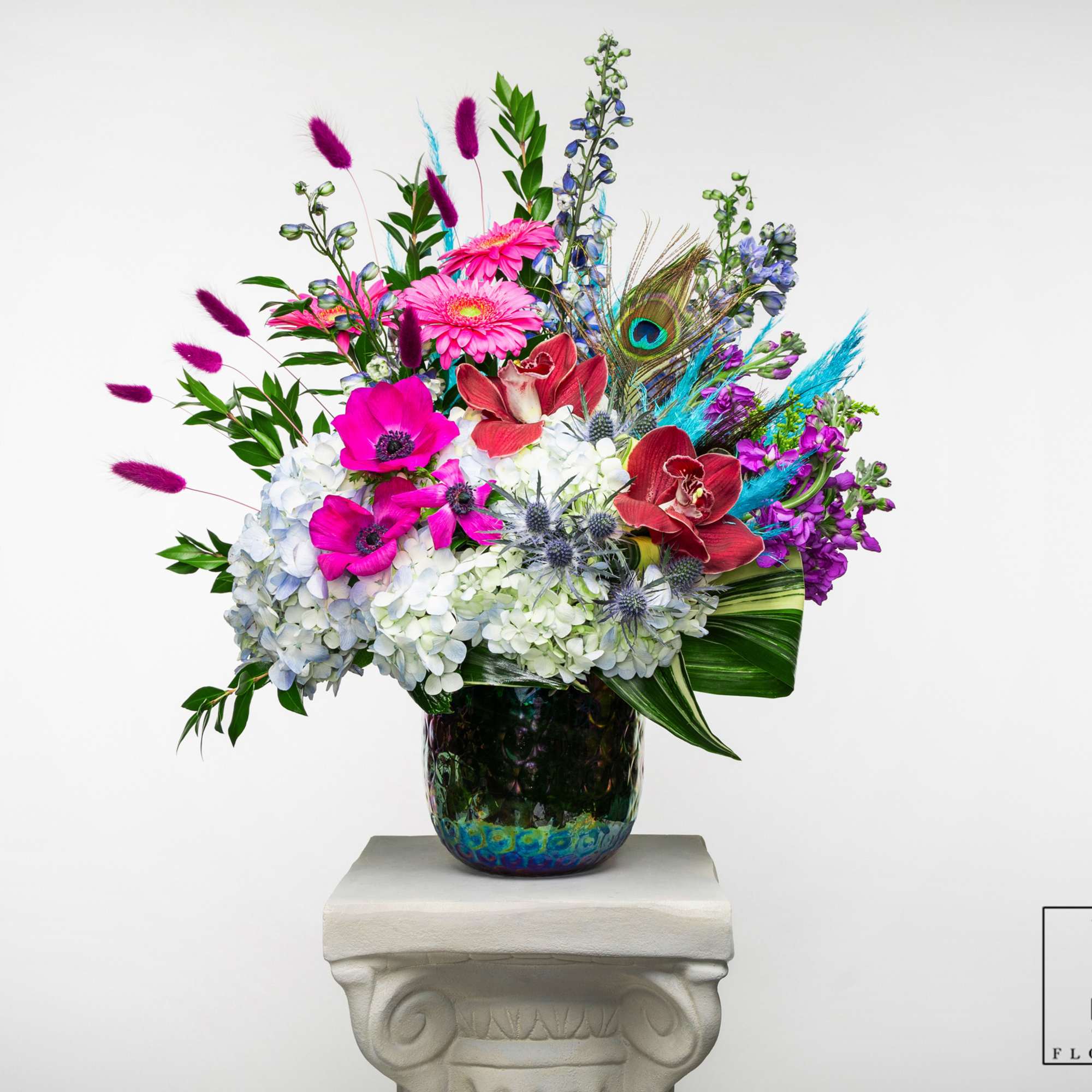 Tall mixed arrangement with white hydrangeas, bright pink blooms, orchids and peacock feather in iridescent vase.