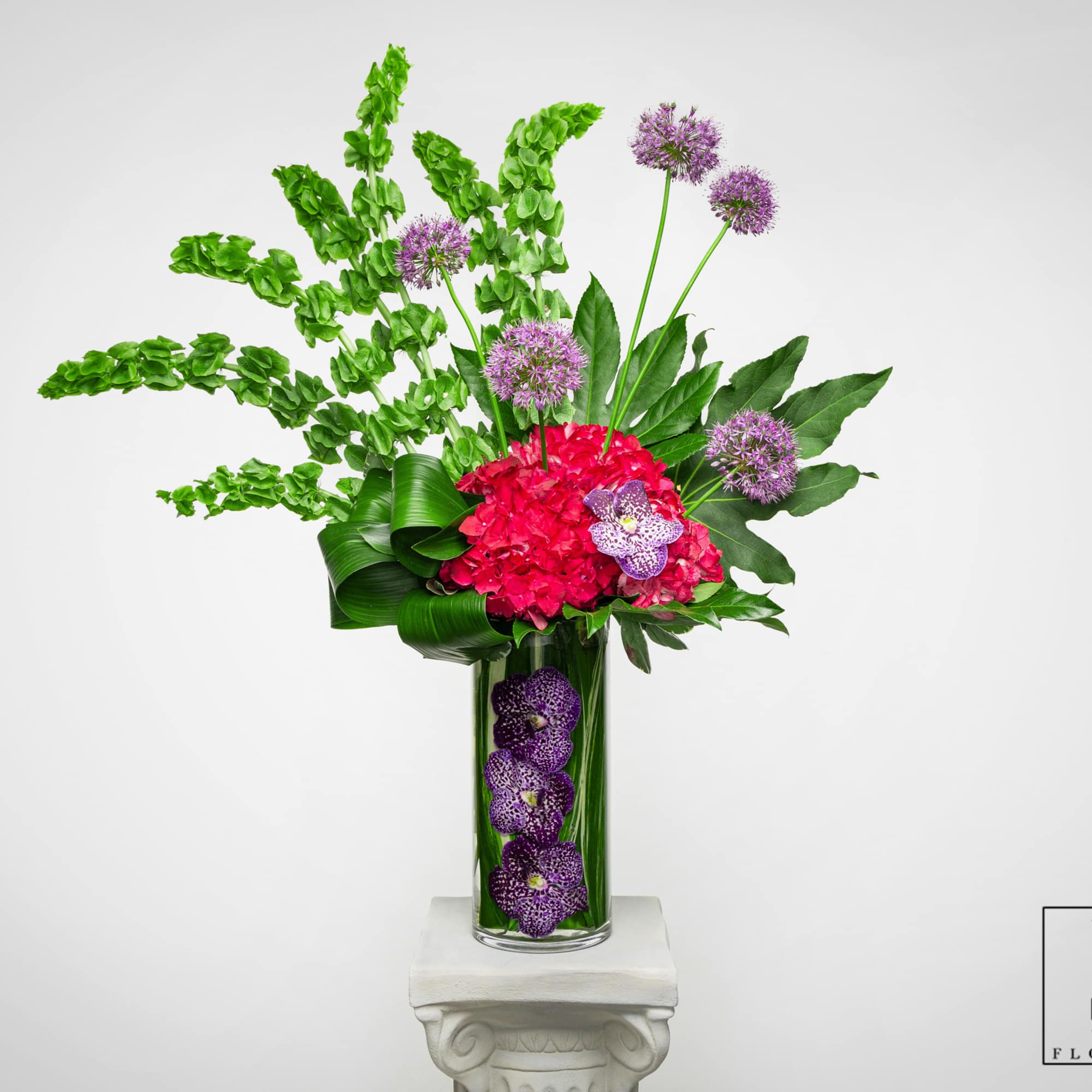 Tall modern arrangement with green bells of Ireland, pink hydrangea and purple orchids in a glass vase