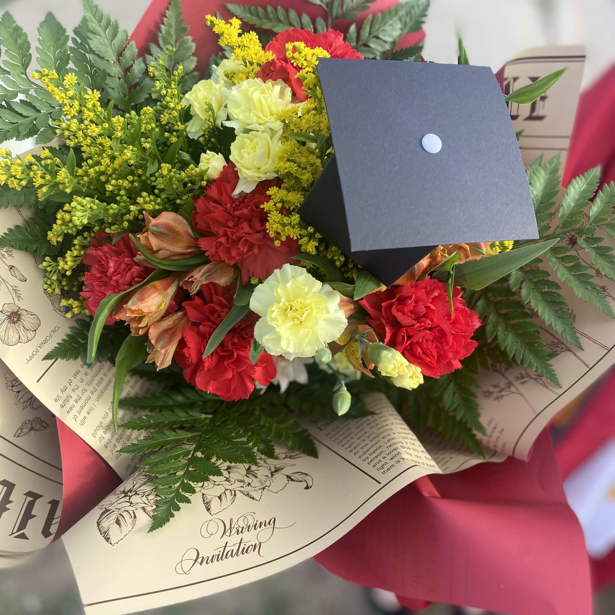 Bouquet of red and pale yellow carnations with greenery and a graduation cap