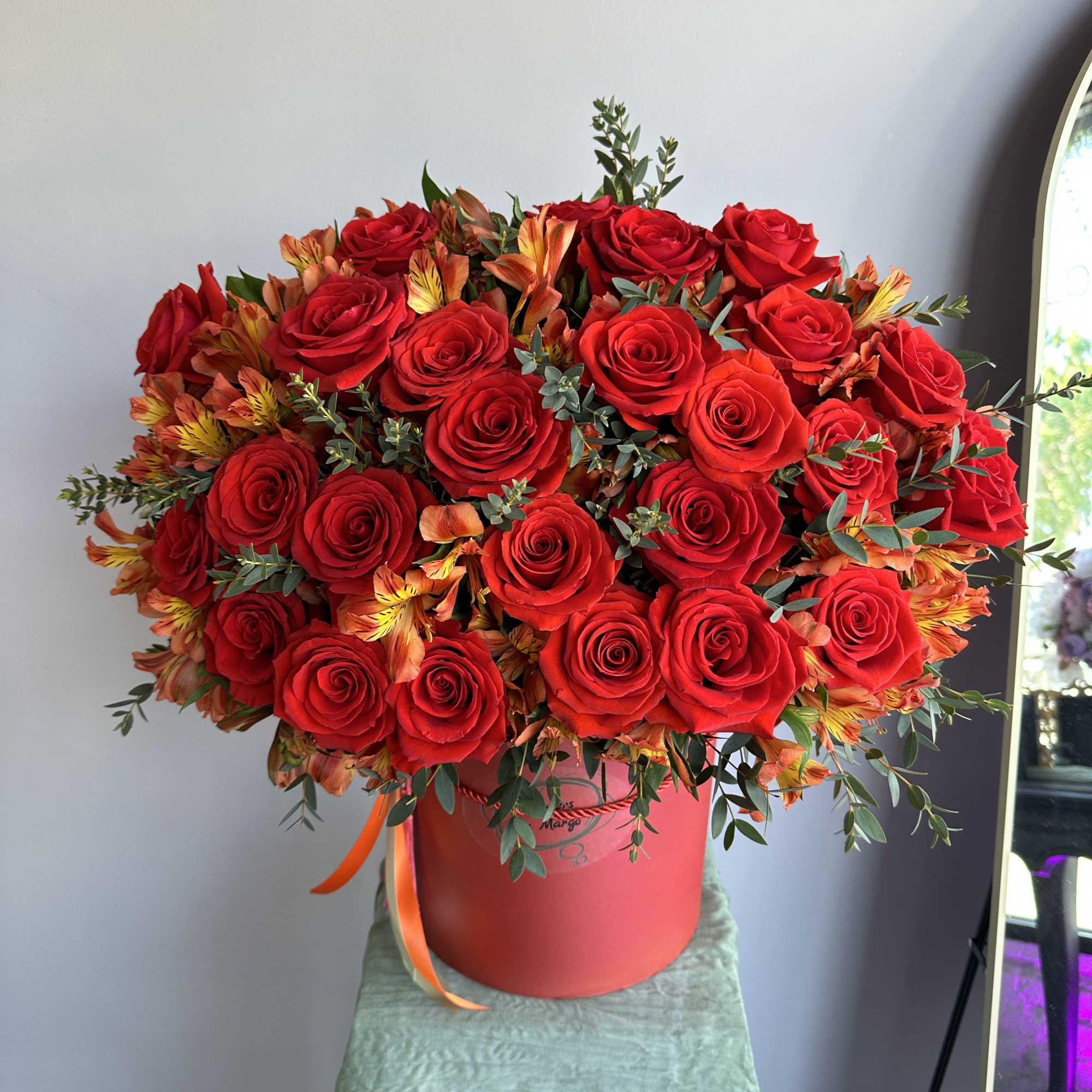 Large arrangement of red roses and orange flowers in a red hatbox container.