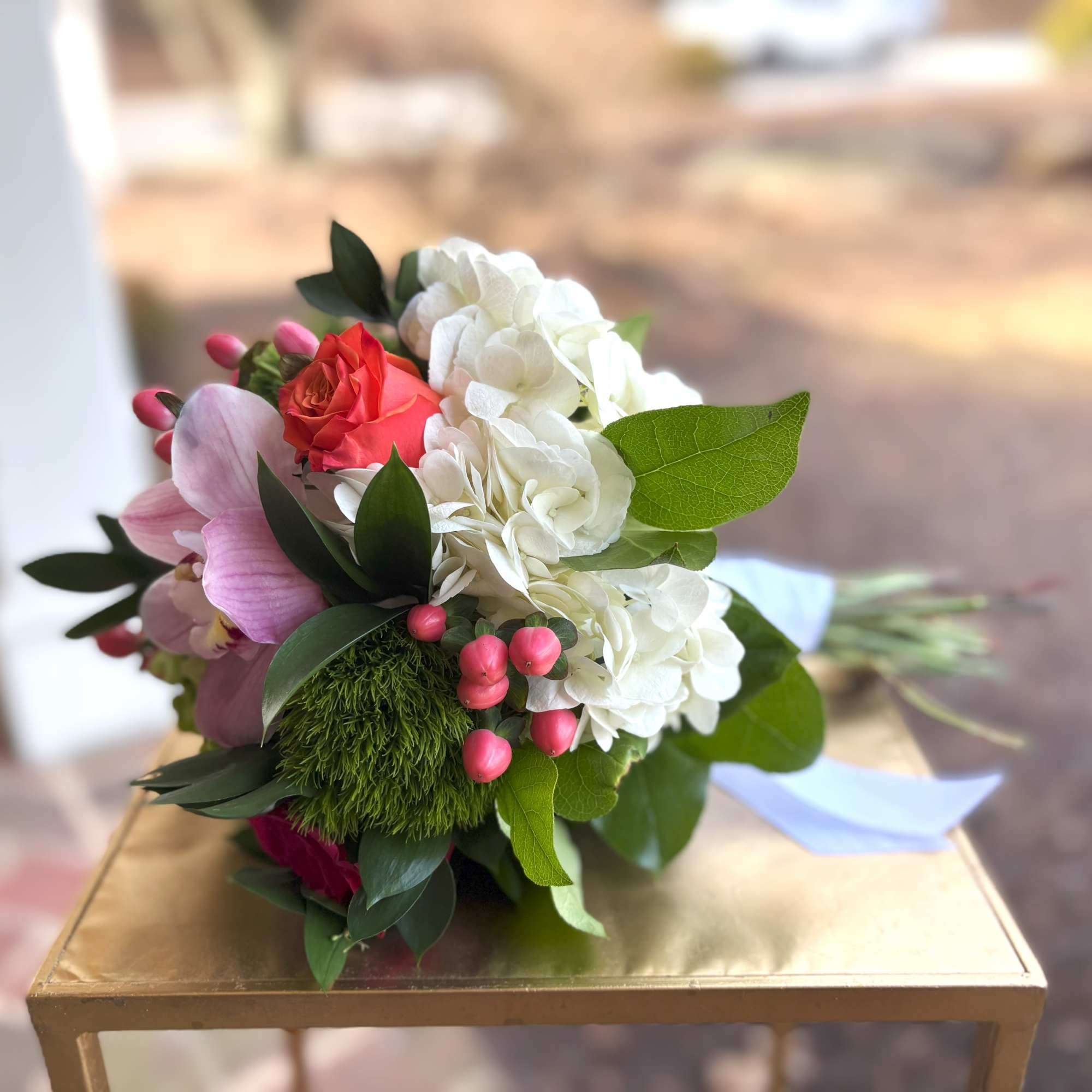 Hand-tied bouquet with white hydrangeas, coral roses, pink orchids, and berries, wrapped with a blue ribbon.