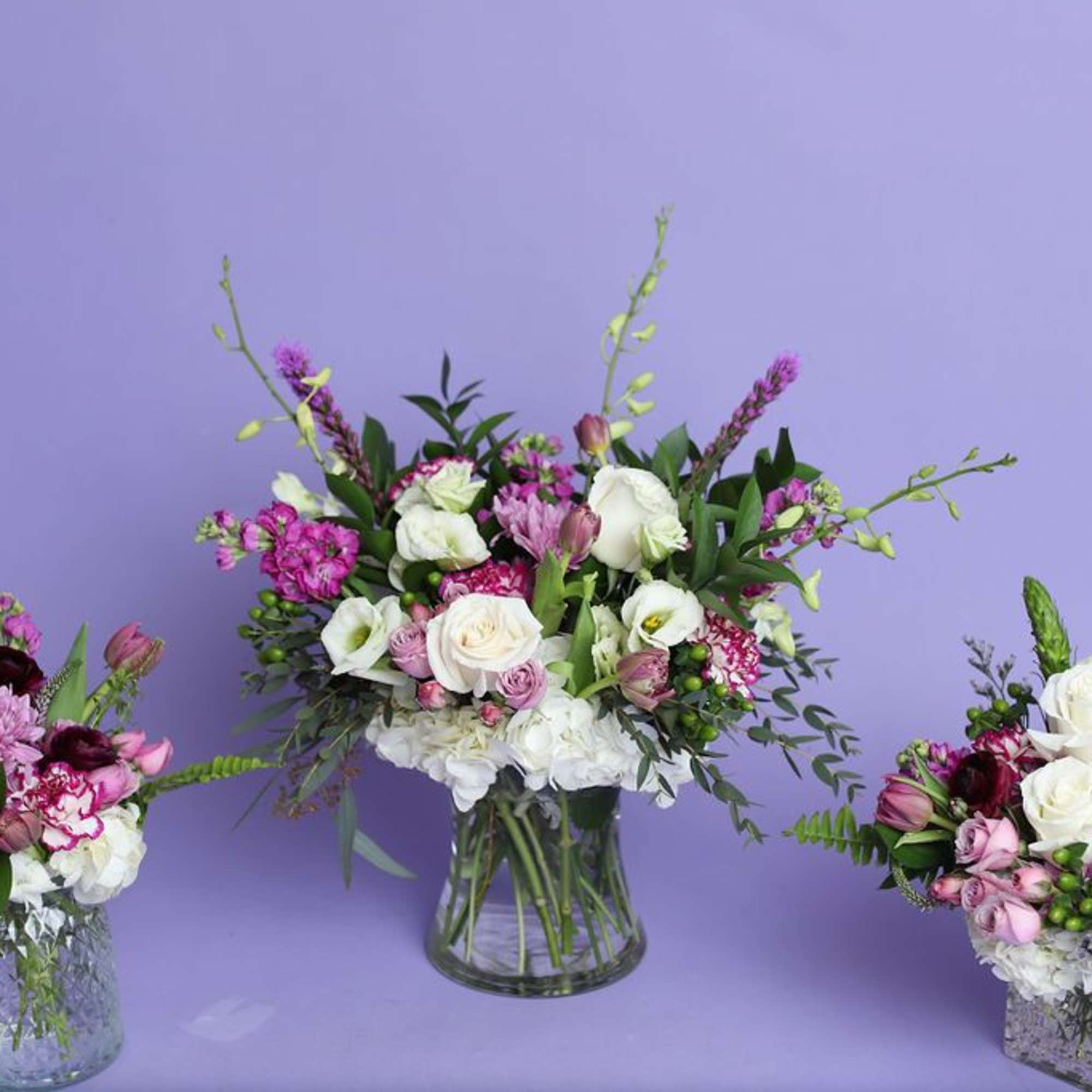 Three mixed floral arrangements in glass vases on a lavender background