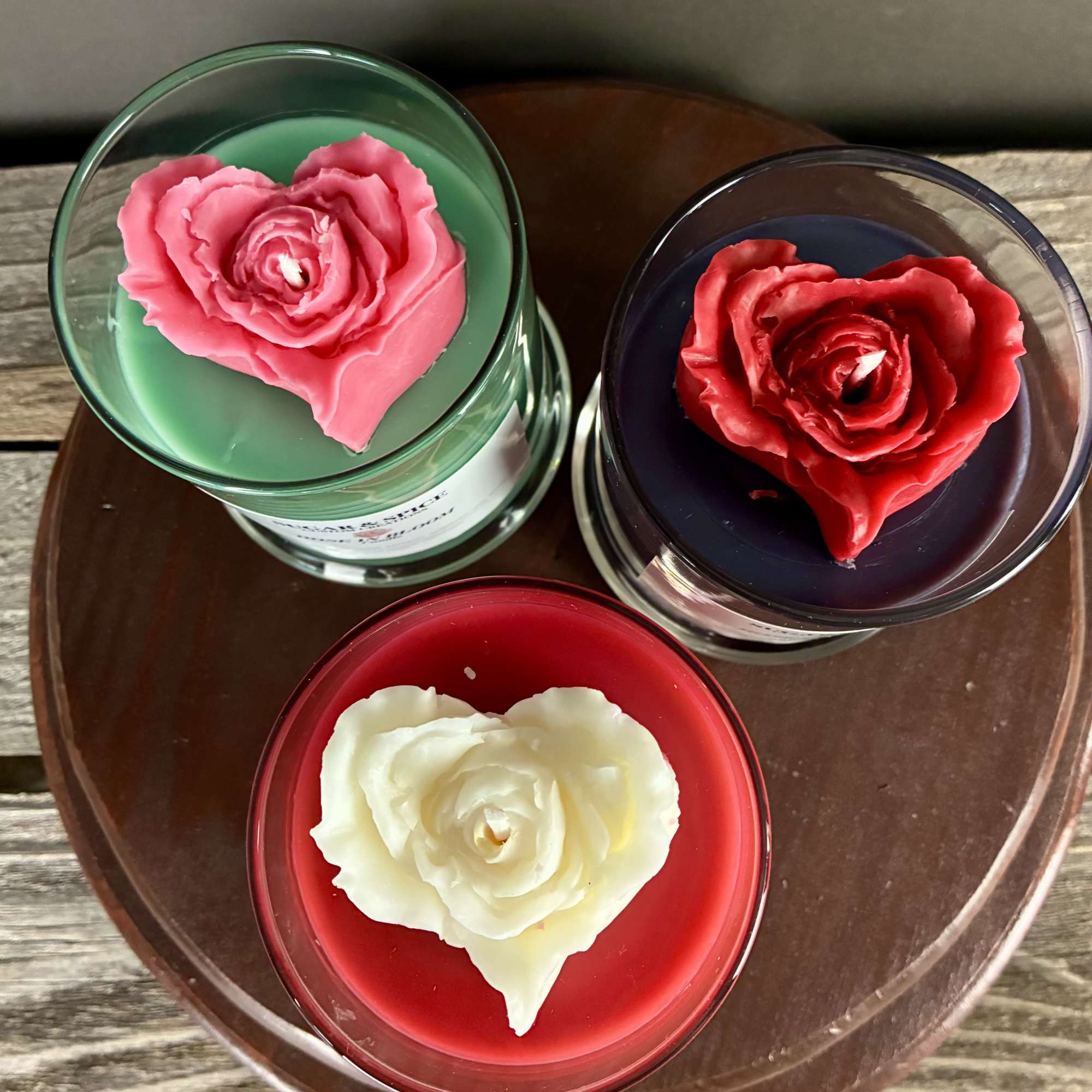 Trio of glass jar candles with heart-shaped rose wax tops in red, pink, and white on a wooden tray