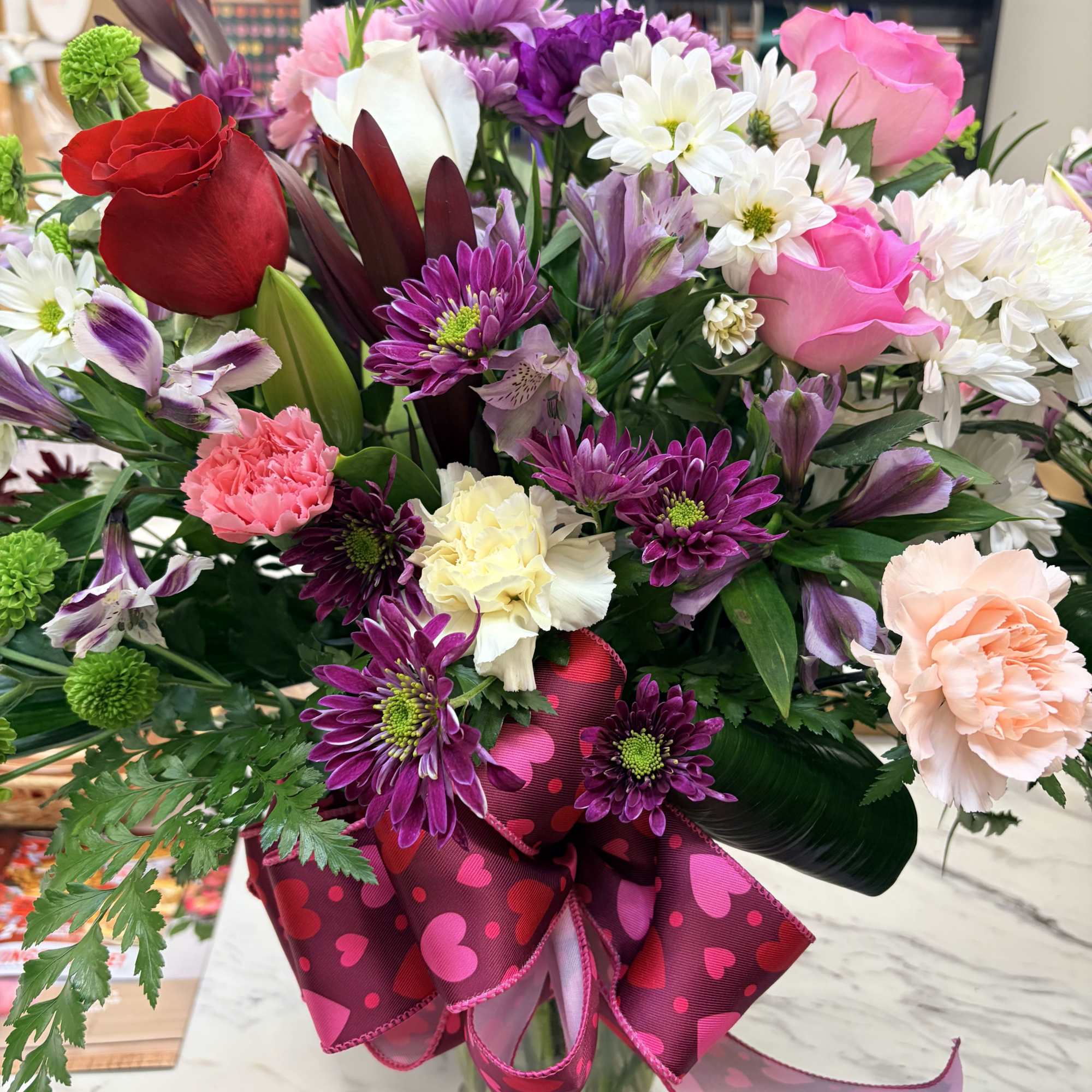 Mixed bouquet of roses, daisies, and carnations in a glass vase with a pink ribbon